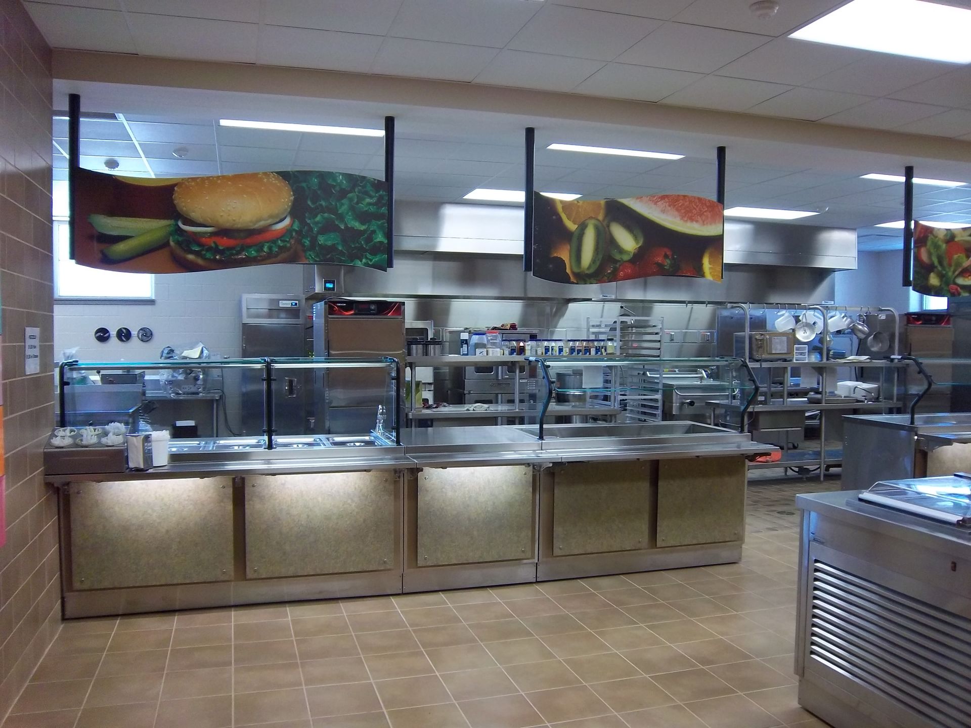 A commercial cafeteria serving line with stainless steel counters and overhead menu boards featuring food images.