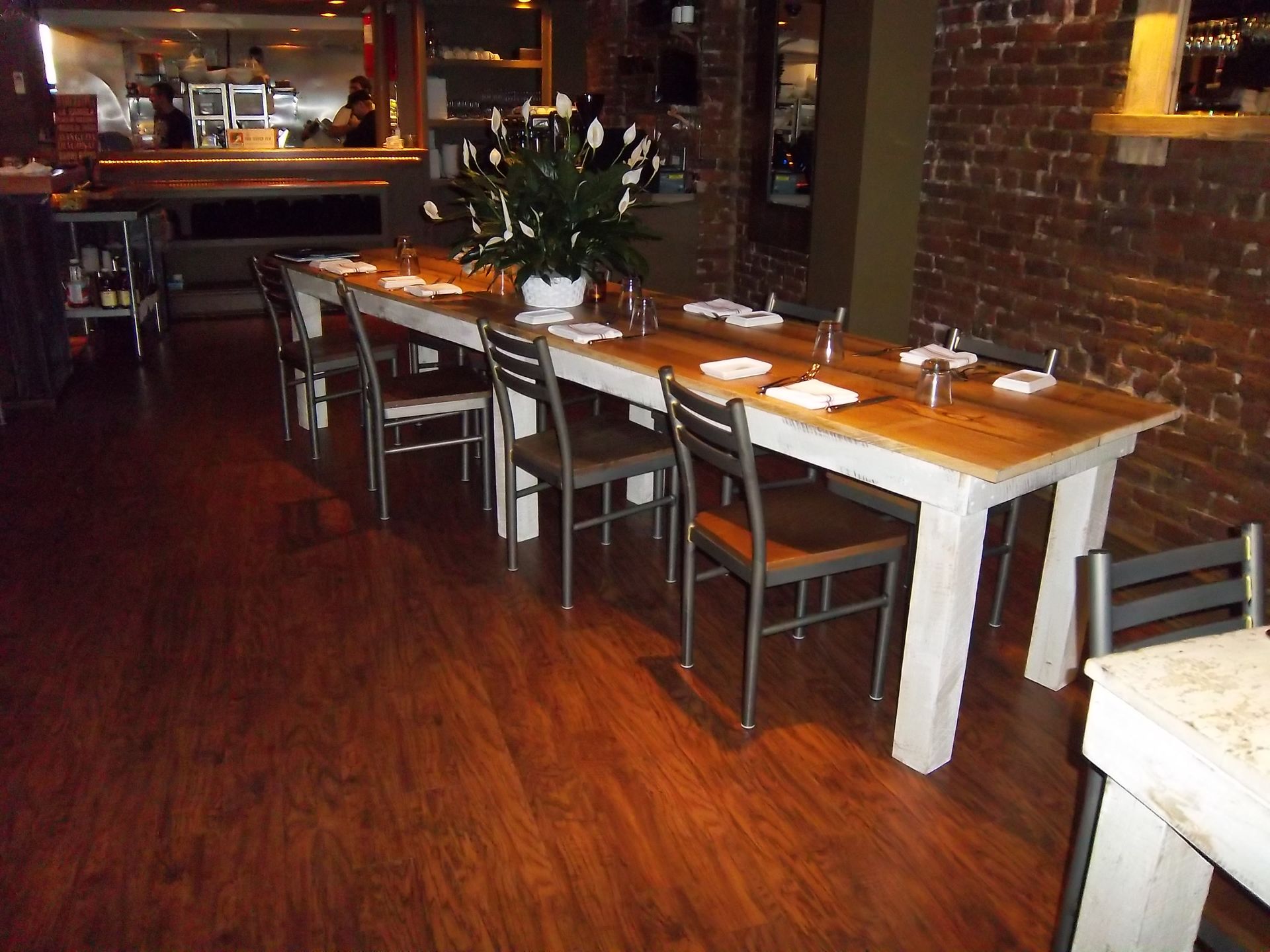 A long wooden dining table with a white base and chairs set in a restaurant with a brick wall and dark wood flooring.