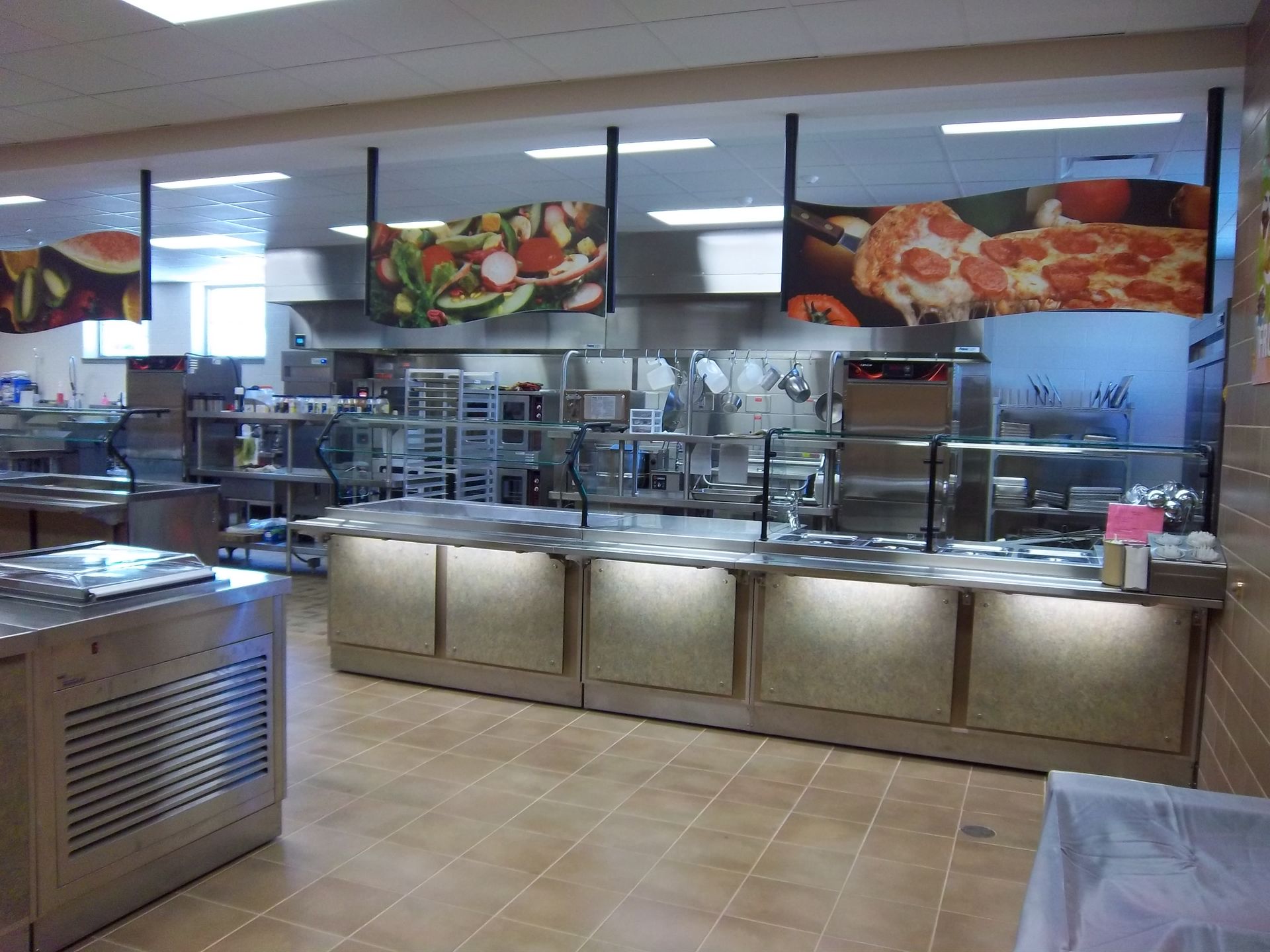 A cafeteria serving area with stainless steel counters and overhead signs featuring images of pizza and fresh salads.