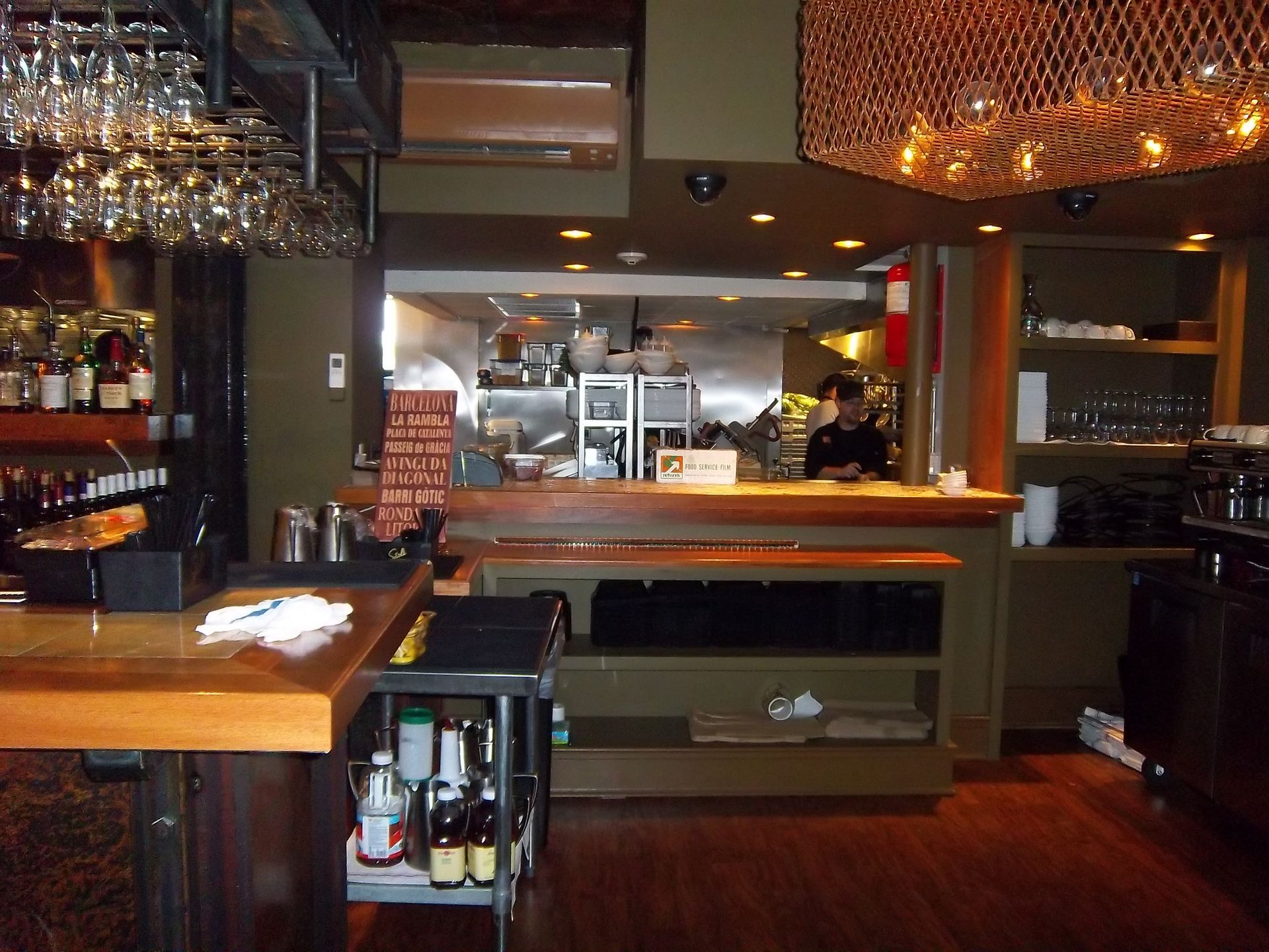 The interior of a bar or restaurant with wooden counters, a staff member in the background, and hanging wine glasses.