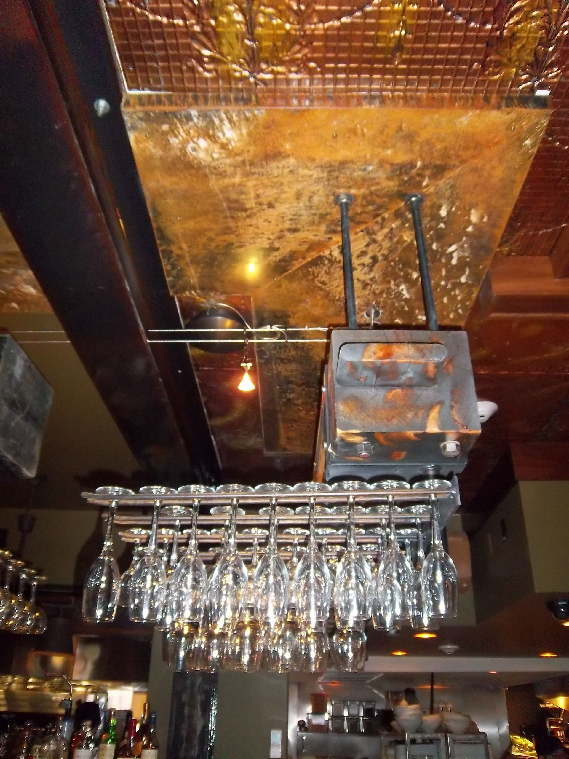 A metal glass rack hanging from a rustic, metallic ceiling, filled with many stemmed glasses in a dim restaurant bar.