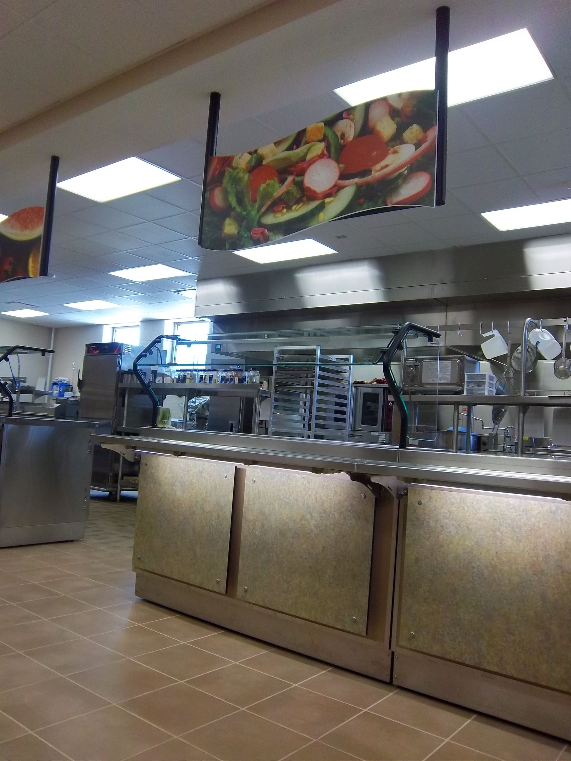 A cafeteria food service line with stainless steel counters and overhead signs featuring colorful salad imagery.