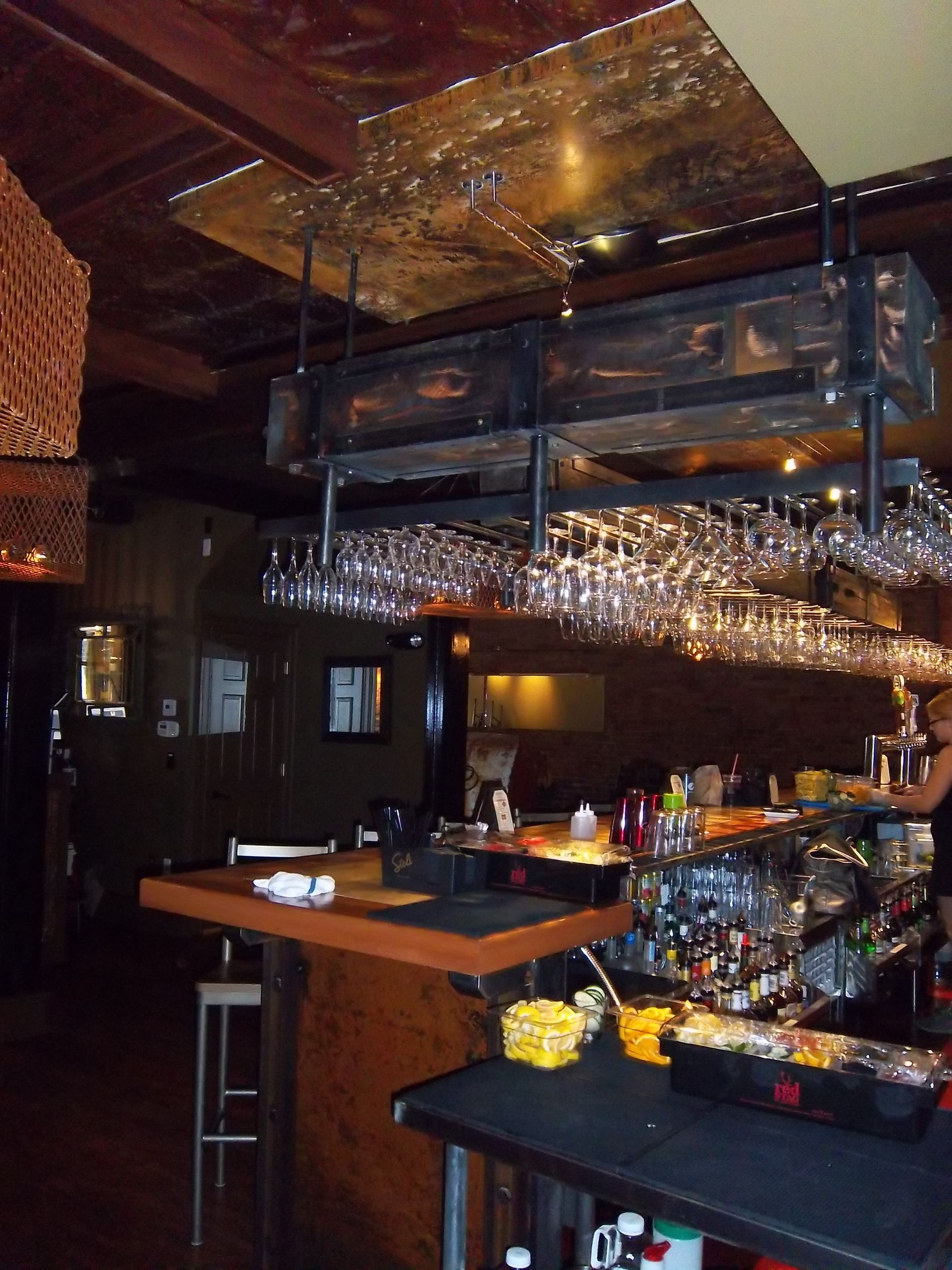 An indoor bar with a wooden counter, a metal overhead glass rack filled with wine glasses, and dim, warm lighting.
