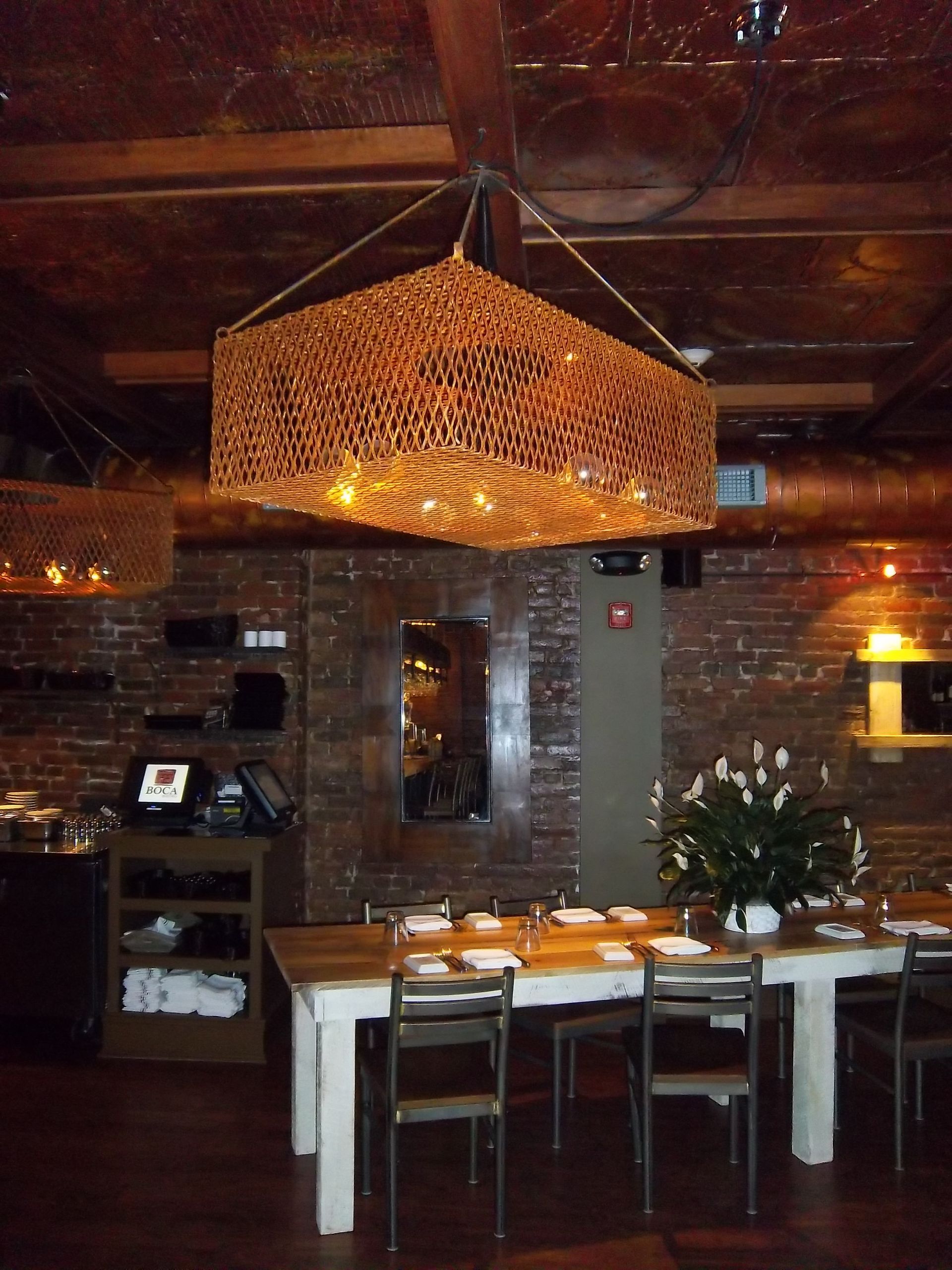A dining area with a rustic wooden table, metal chairs, exposed brick walls, and two large, rectangular mesh pendants.