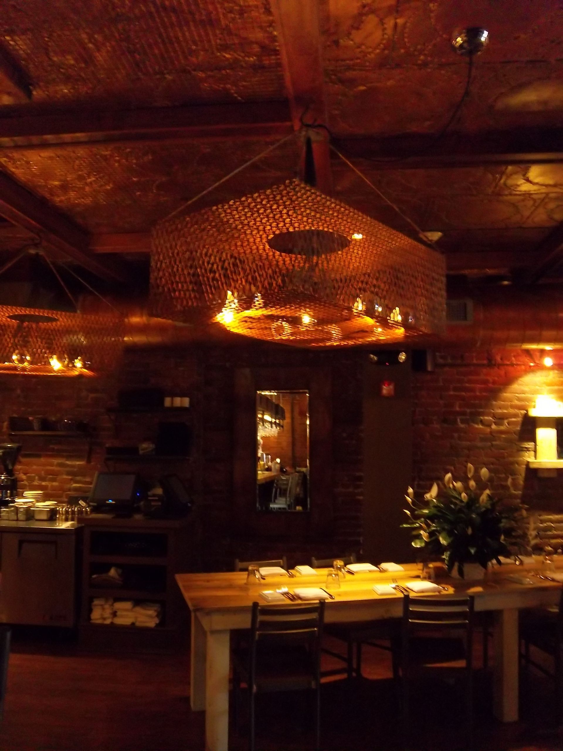 Warmly lit rustic restaurant interior with brick walls, a long dining table, and large, textured woven light fixtures.