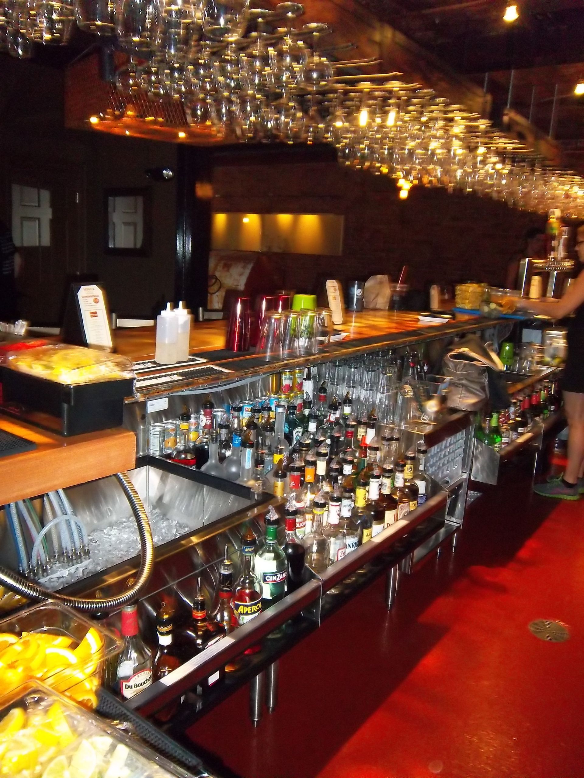 A well-stocked bar counter with rows of bottles, hanging glassware, and a bright red floor.