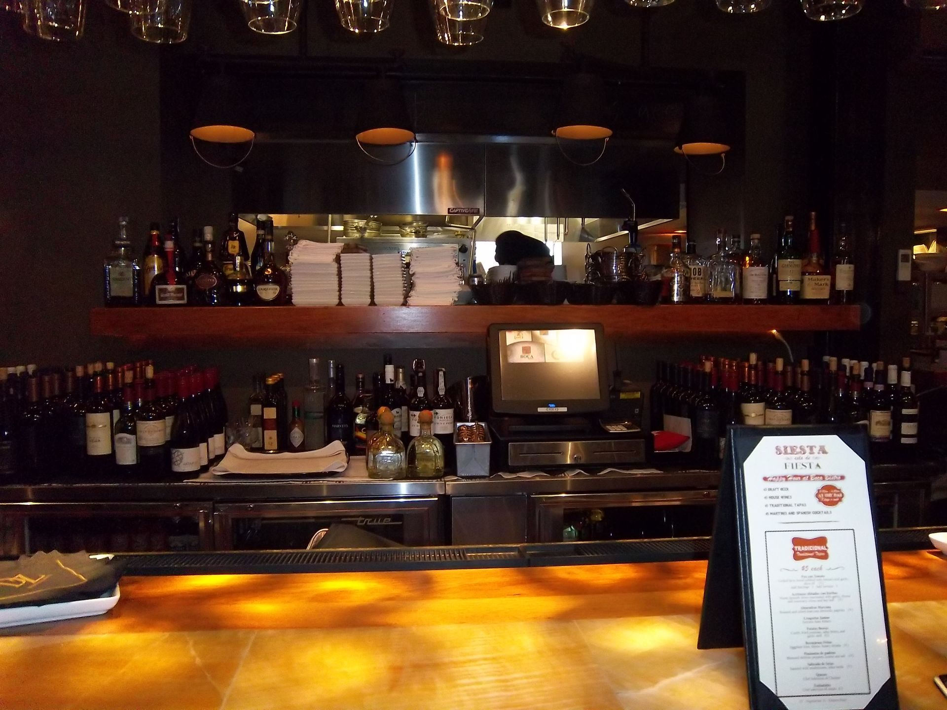 An amber-lit bar counter with a menu stand in the foreground and shelves filled with wine bottles in the background.