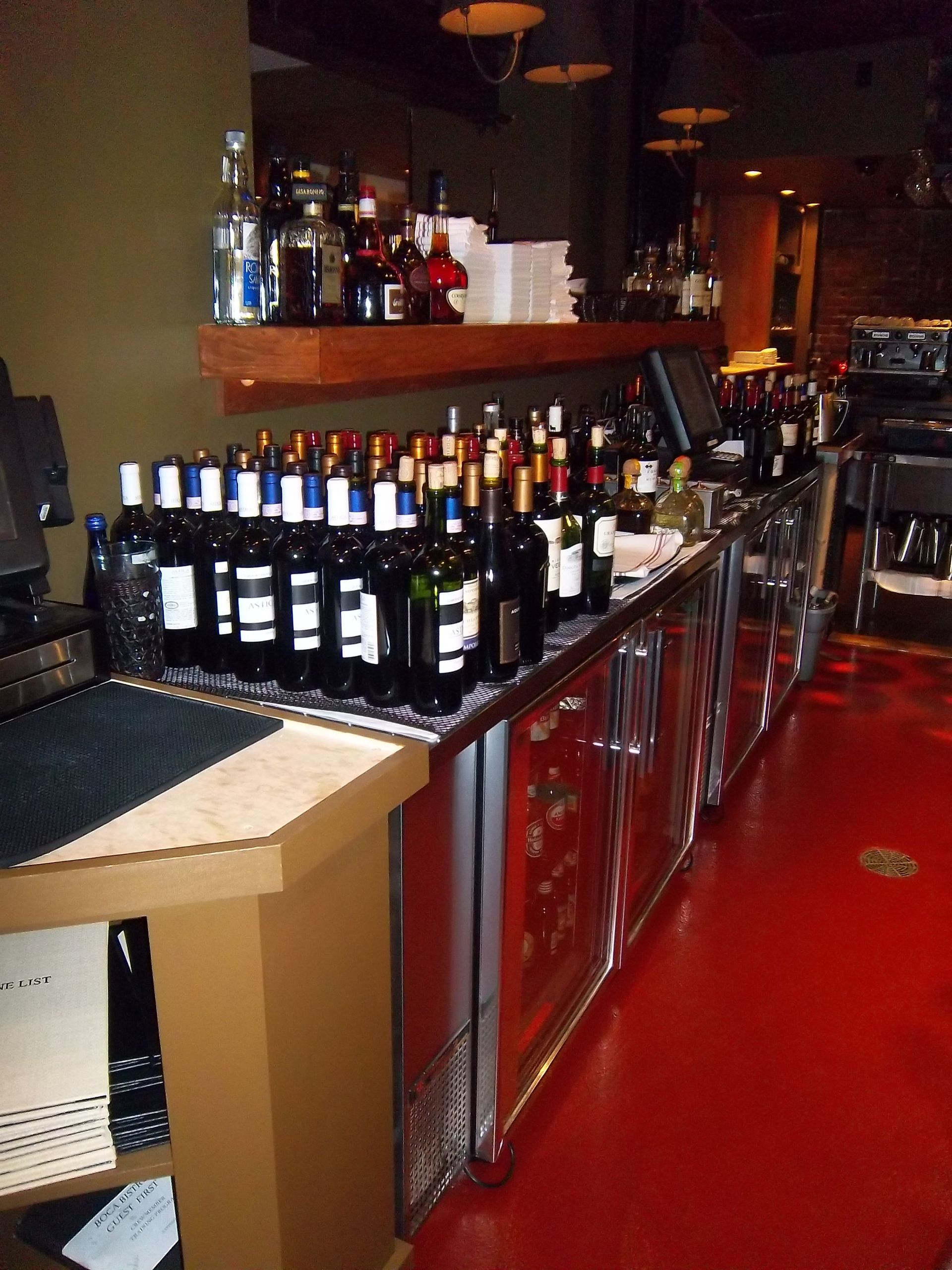 A bar counter with rows of wine bottles stored on top of refrigerated cabinets against a wall, with a red floor.