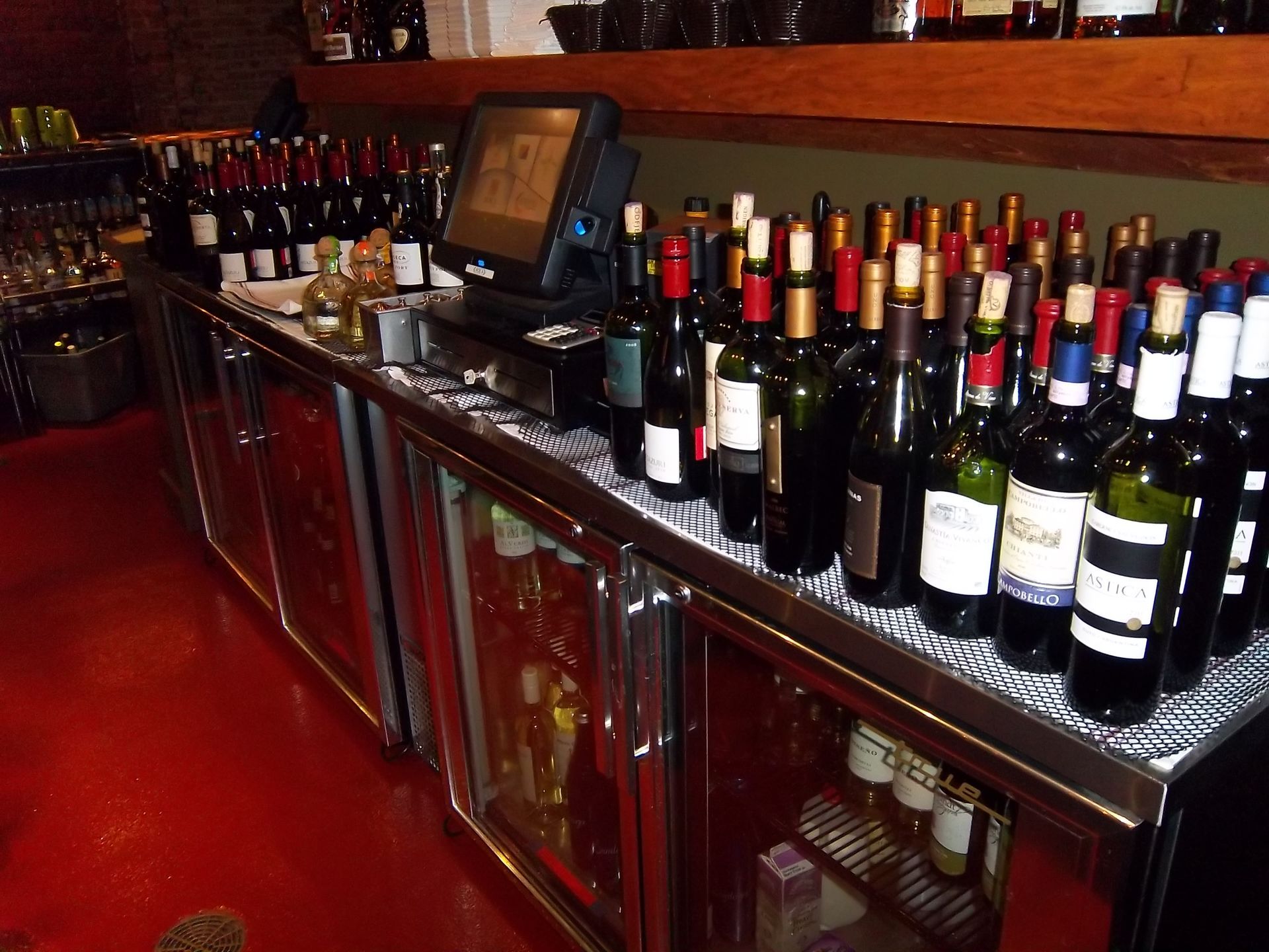 A bar counter with multiple wine bottles arranged in front of a POS system, with glass-front coolers below.