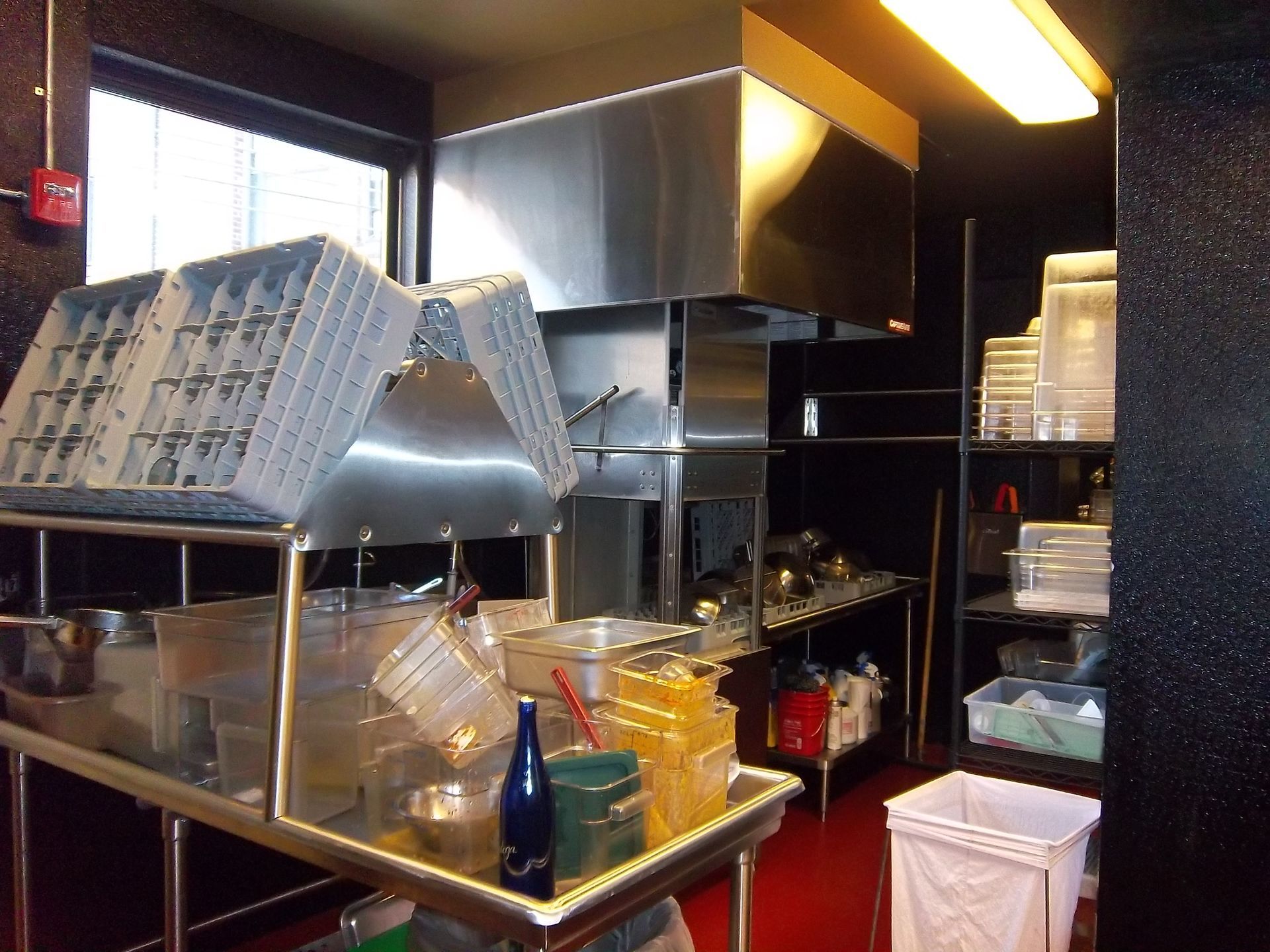 A commercial kitchen with stainless steel prep tables, shelving units with plastic containers, and a large metal hood.