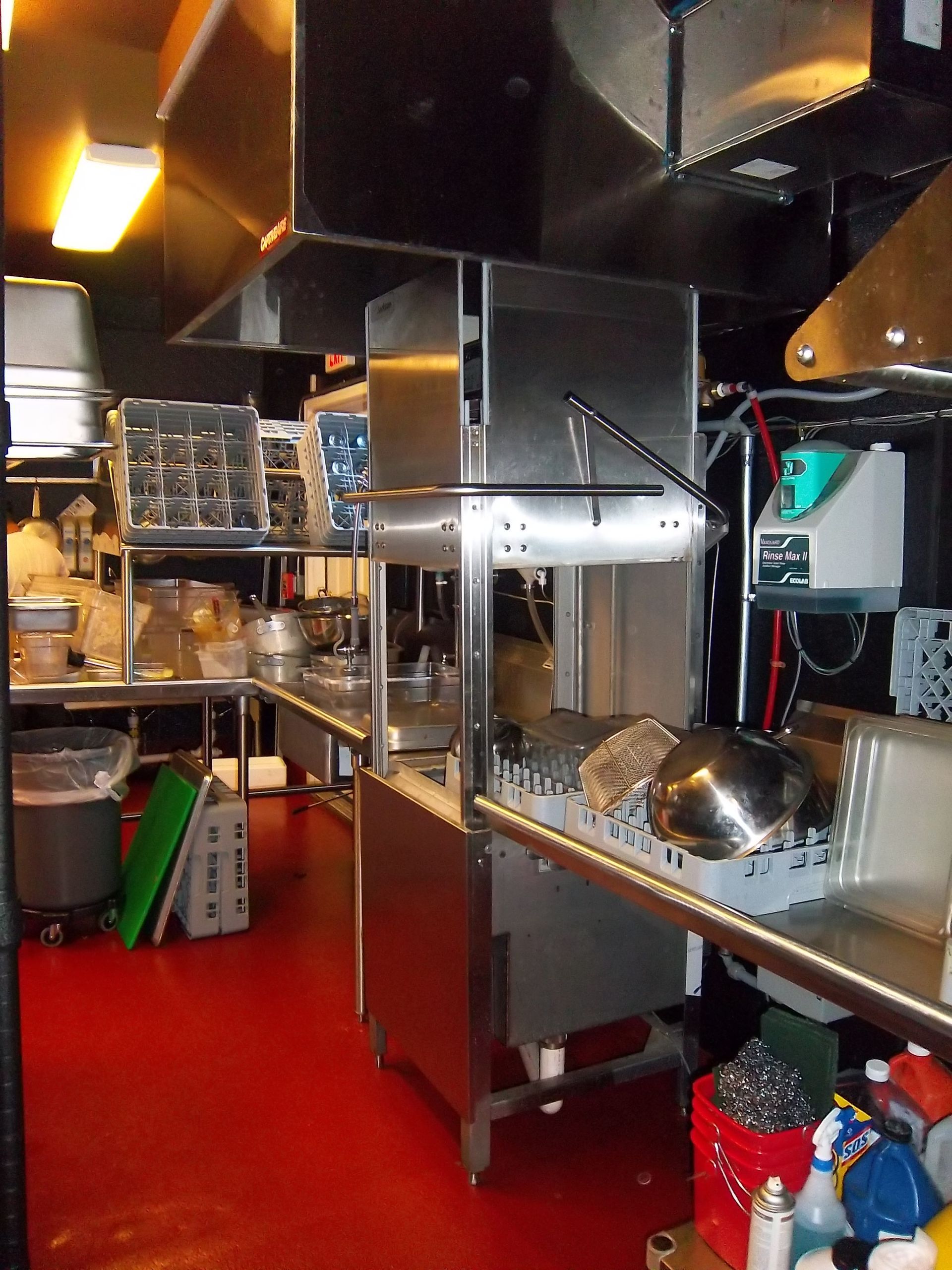 A commercial kitchen featuring a stainless steel dishwasher station, metal prep tables, and a bright red floor.