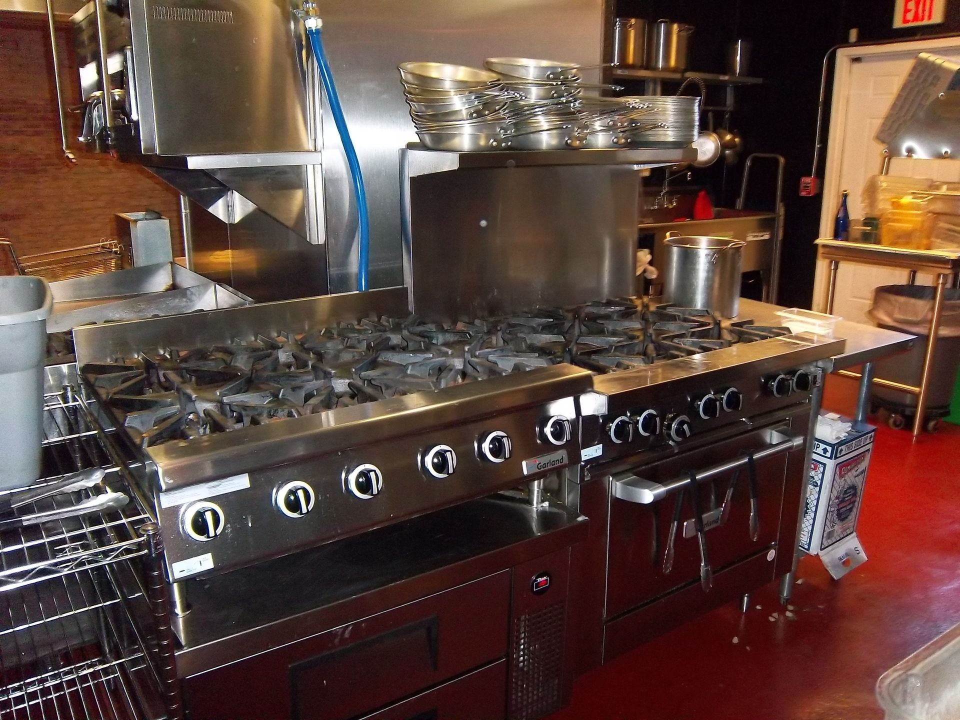 A stainless-steel commercial stove and oven range with burners and knobs sits in a professional kitchen with red flooring.