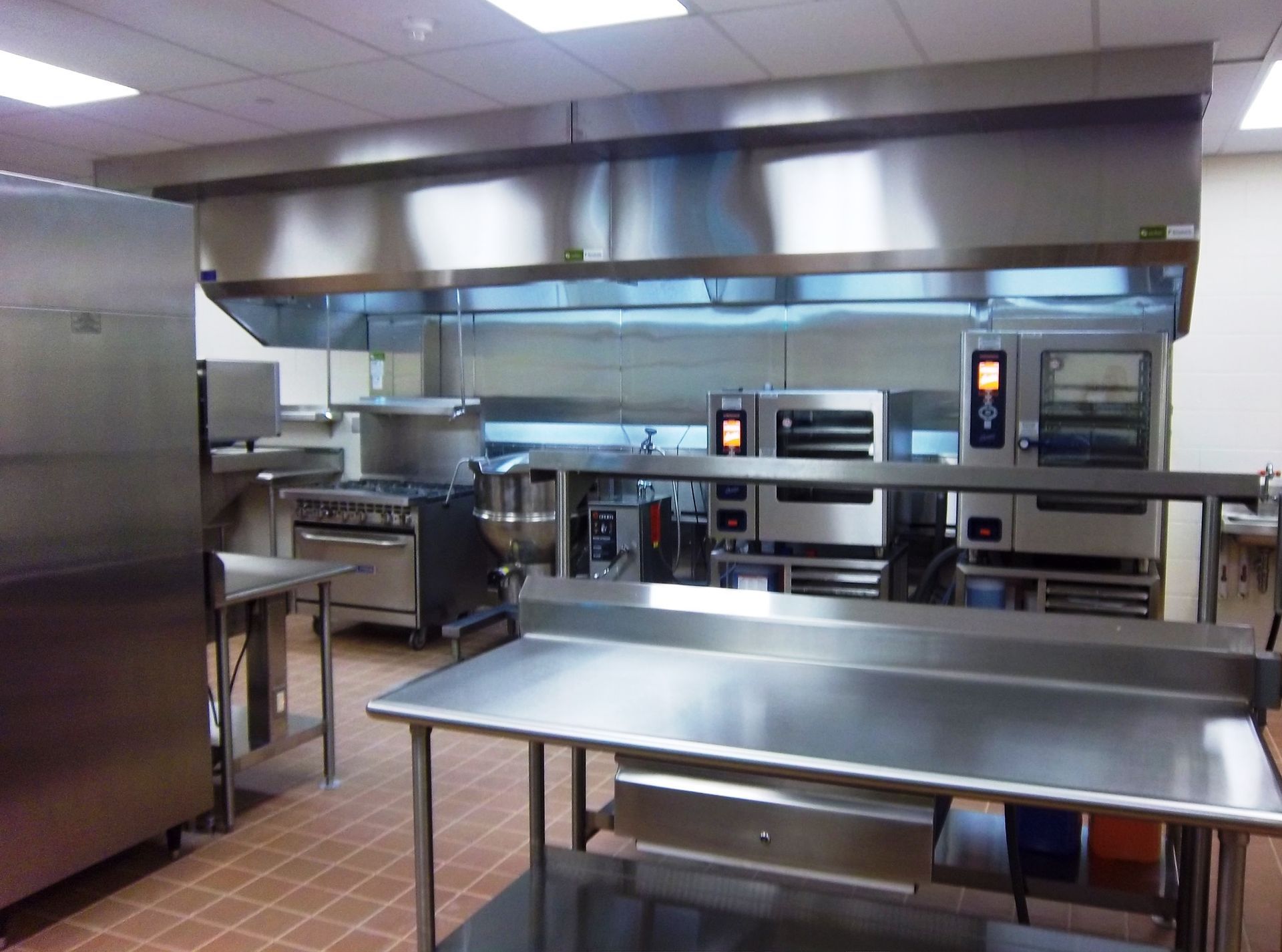 Commercial kitchen featuring stainless steel prep tables, ovens, and a large ventilation hood over the cooking area.
