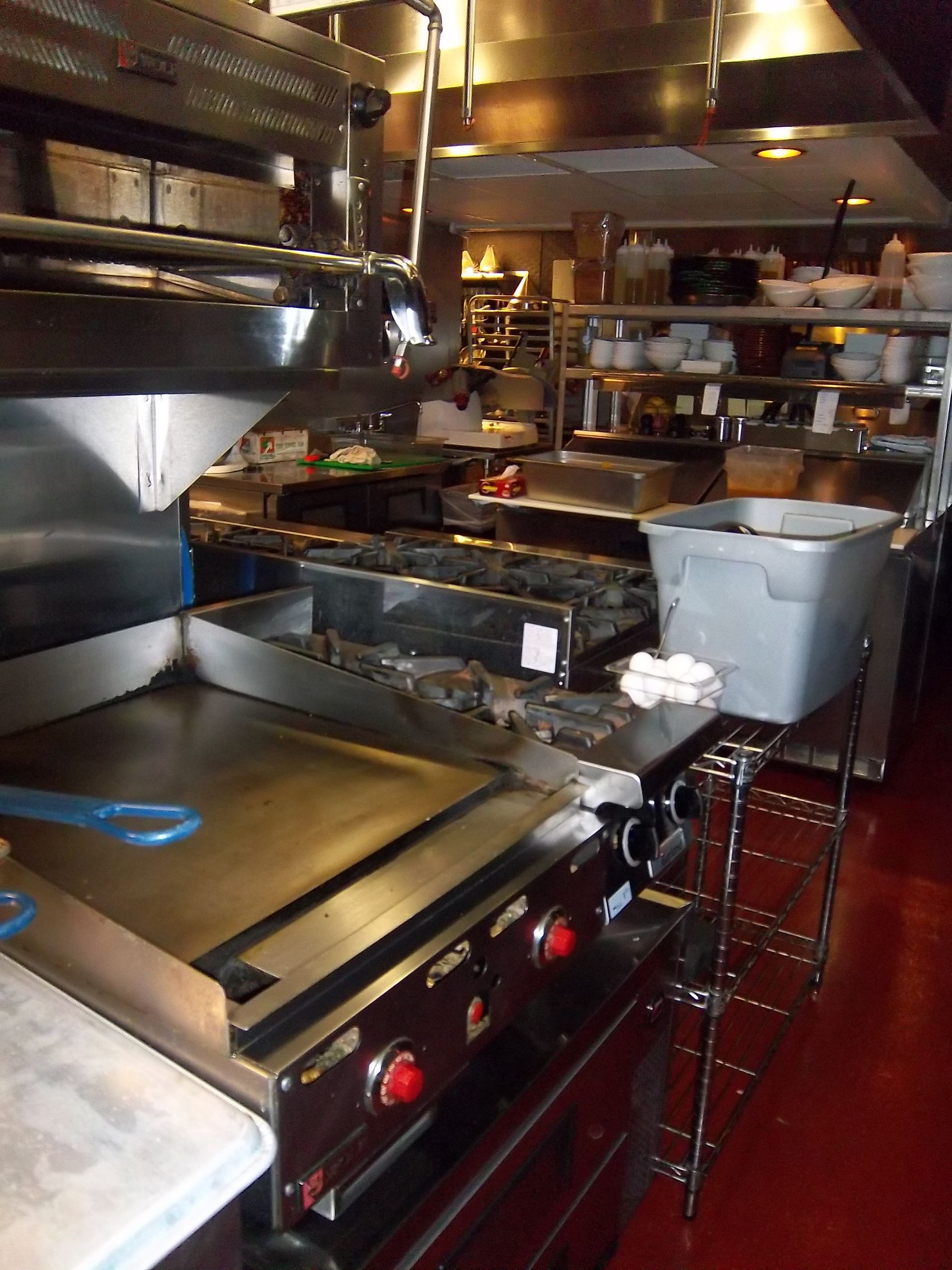 A commercial kitchen featuring a stainless steel flat-top grill in the foreground, shelving units, and a storage bin.