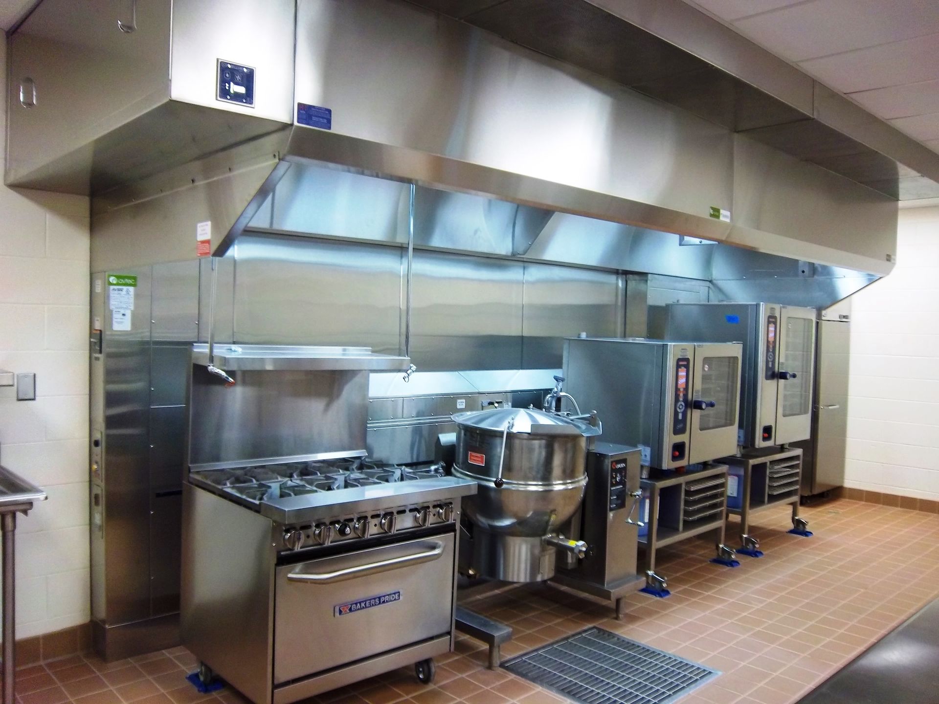 Commercial kitchen featuring stainless steel appliances, including a range, kettle, and ovens, under a large range hood.