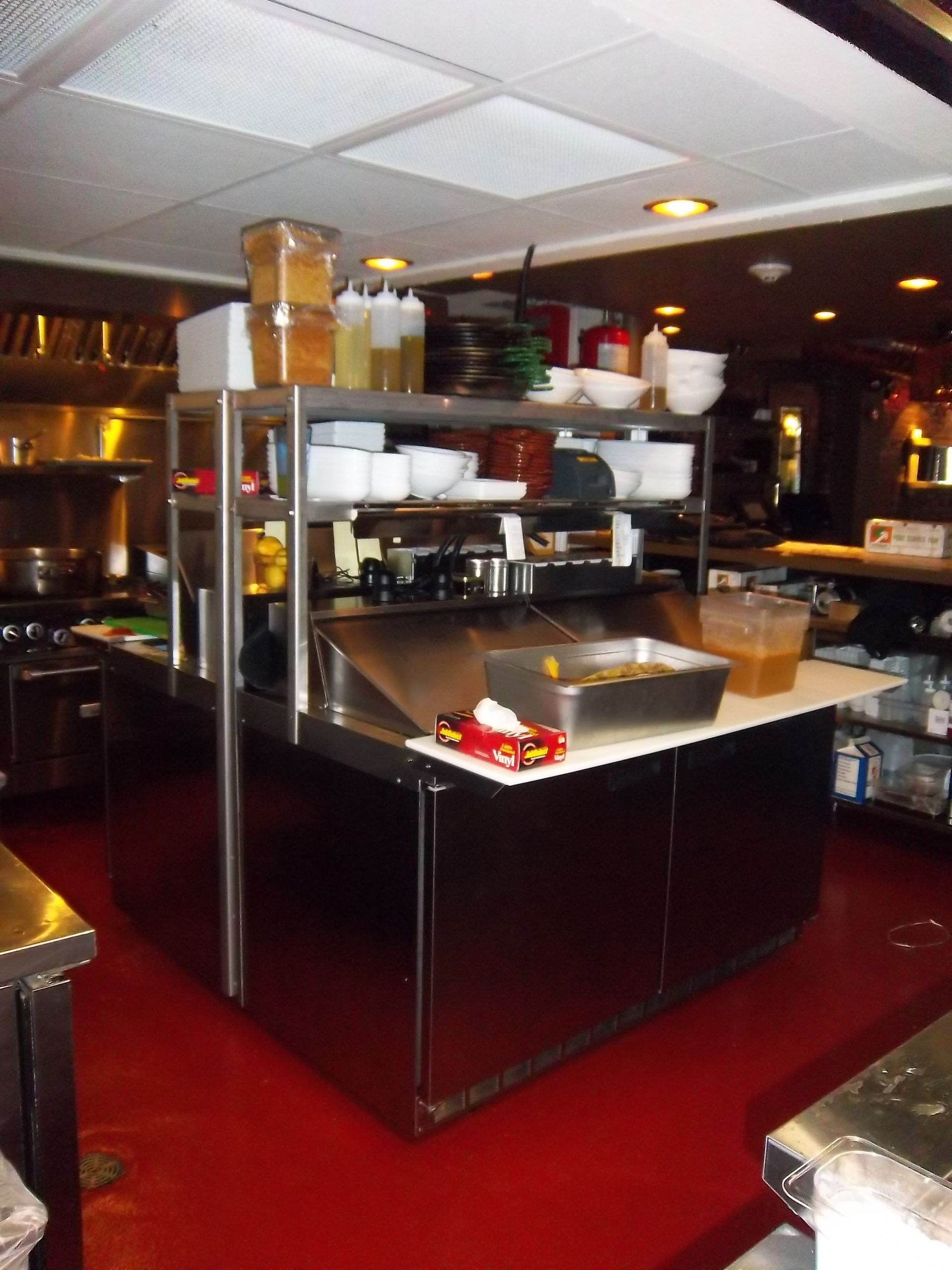 A commercial kitchen station with a stainless steel prep table, shelves holding bowls and bottles, and red flooring.
