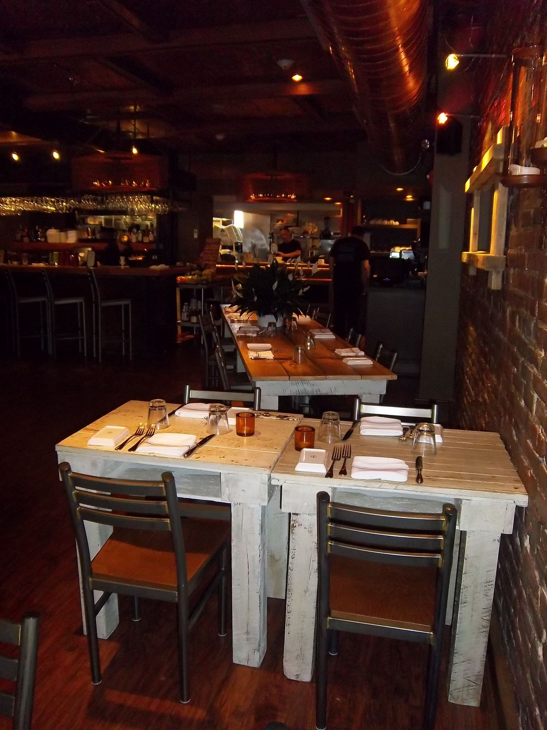 A dimly lit restaurant interior with rustic, white-painted wooden tables set for dining and a bar in the background.
