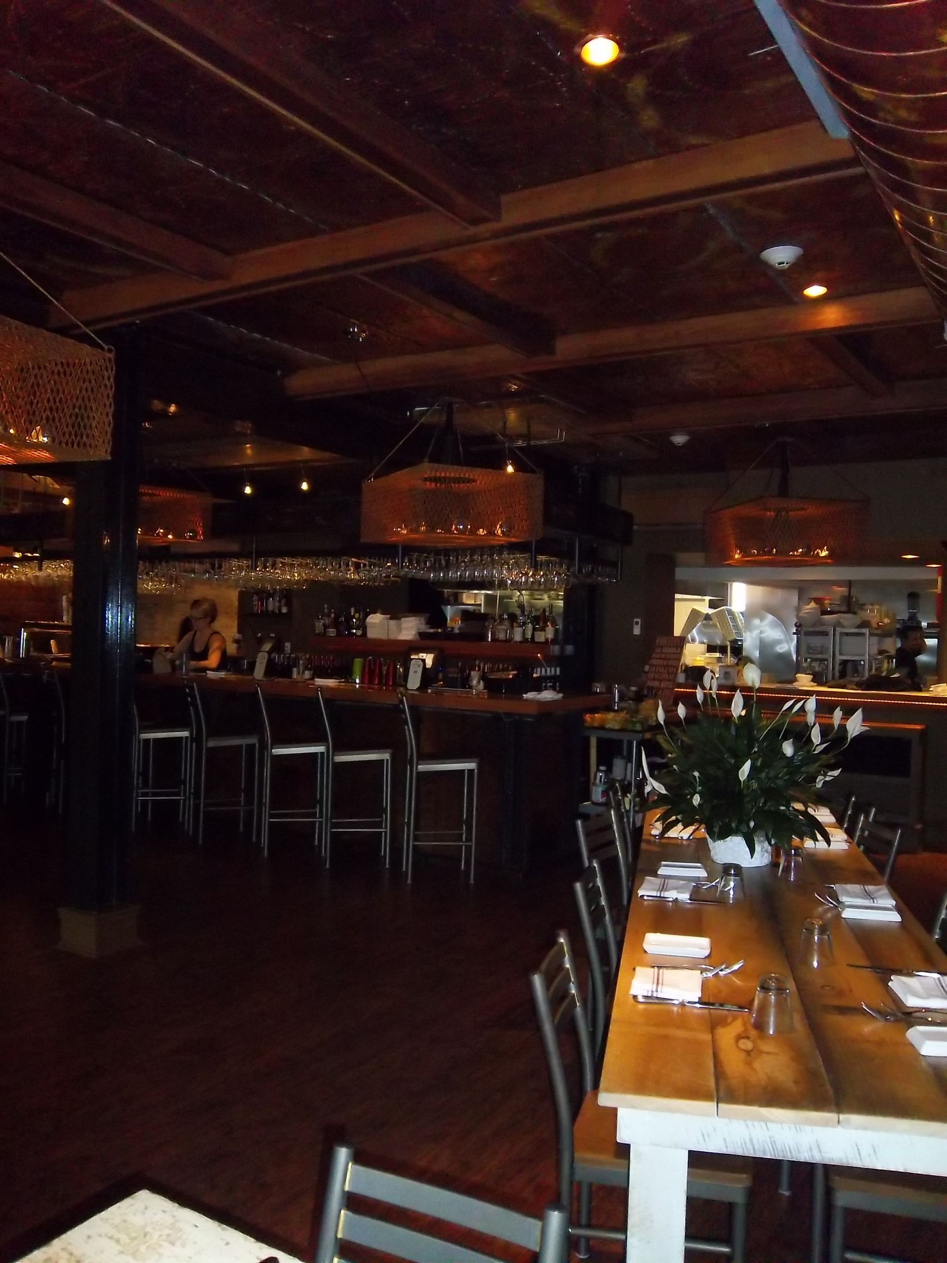 Interior of a rustic restaurant with a wooden long table in the foreground and a bar with stools in the background.