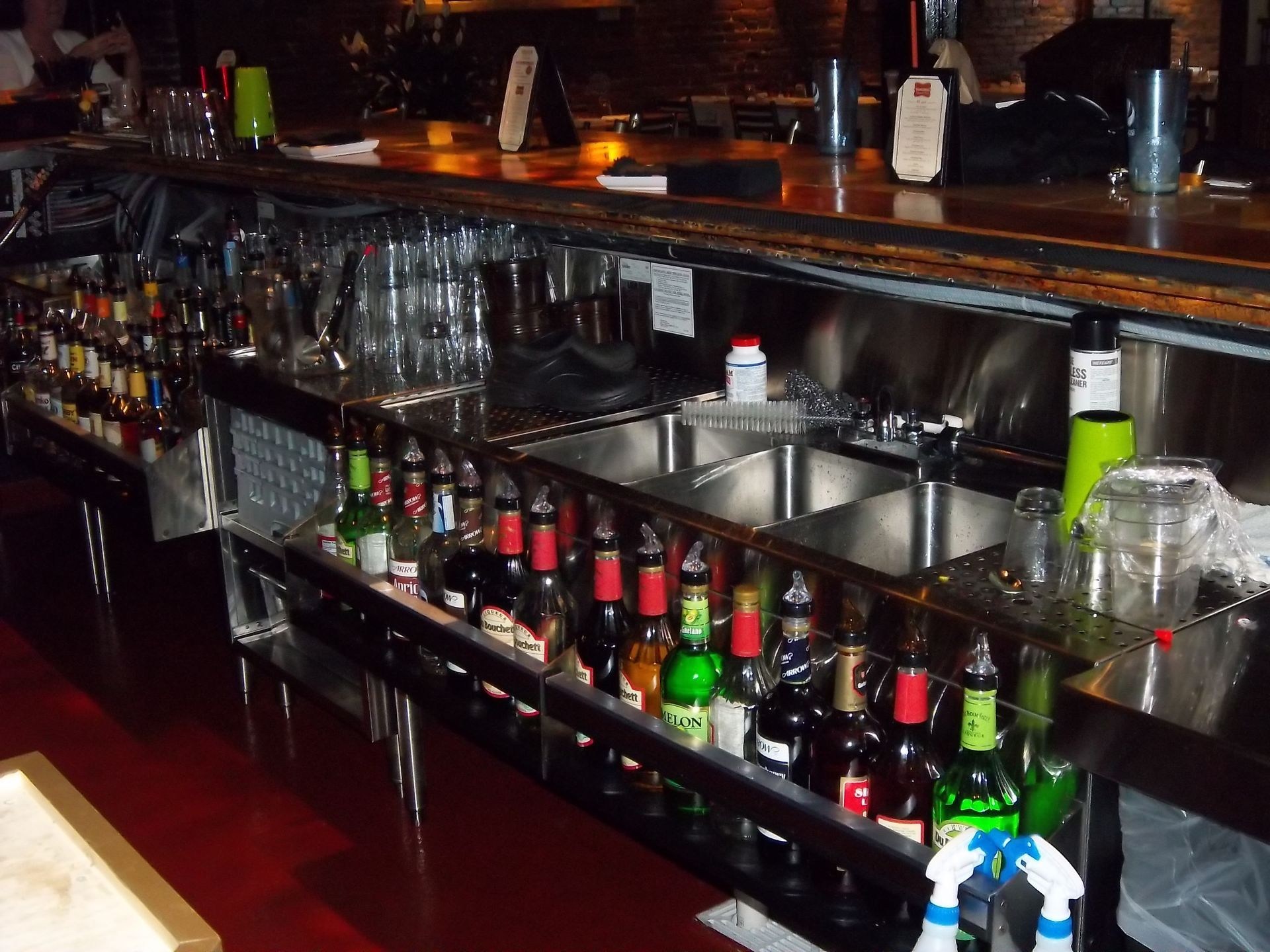 A bar counter featuring shelves stocked with various liquor bottles, a stainless steel sink, and glassware.