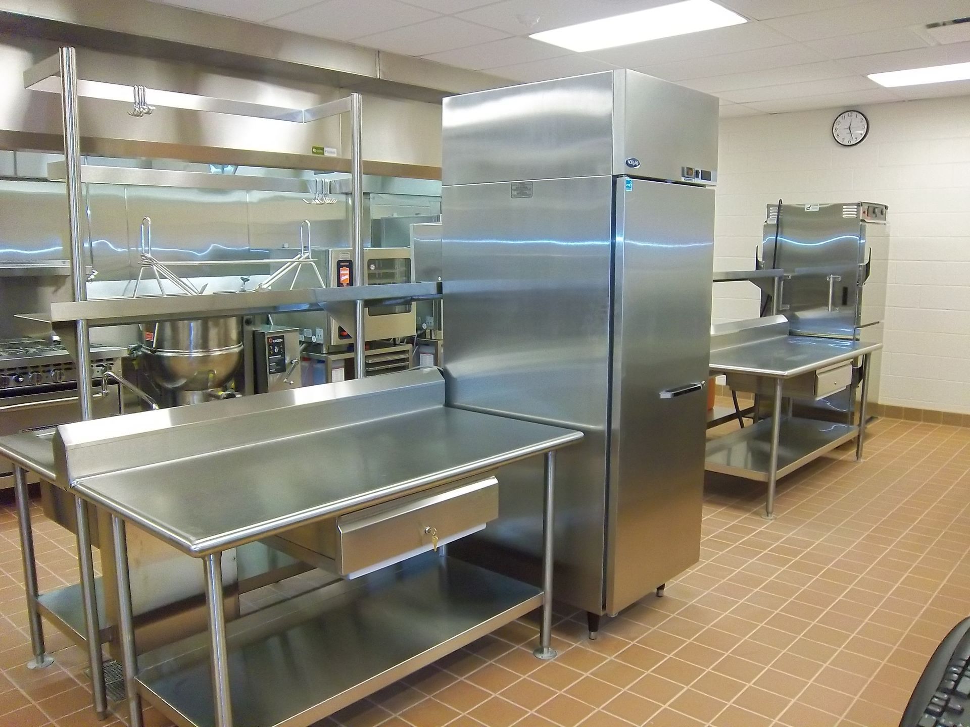 A commercial kitchen featuring stainless steel tables, a tall refrigerator, and shelving on a tiled floor.