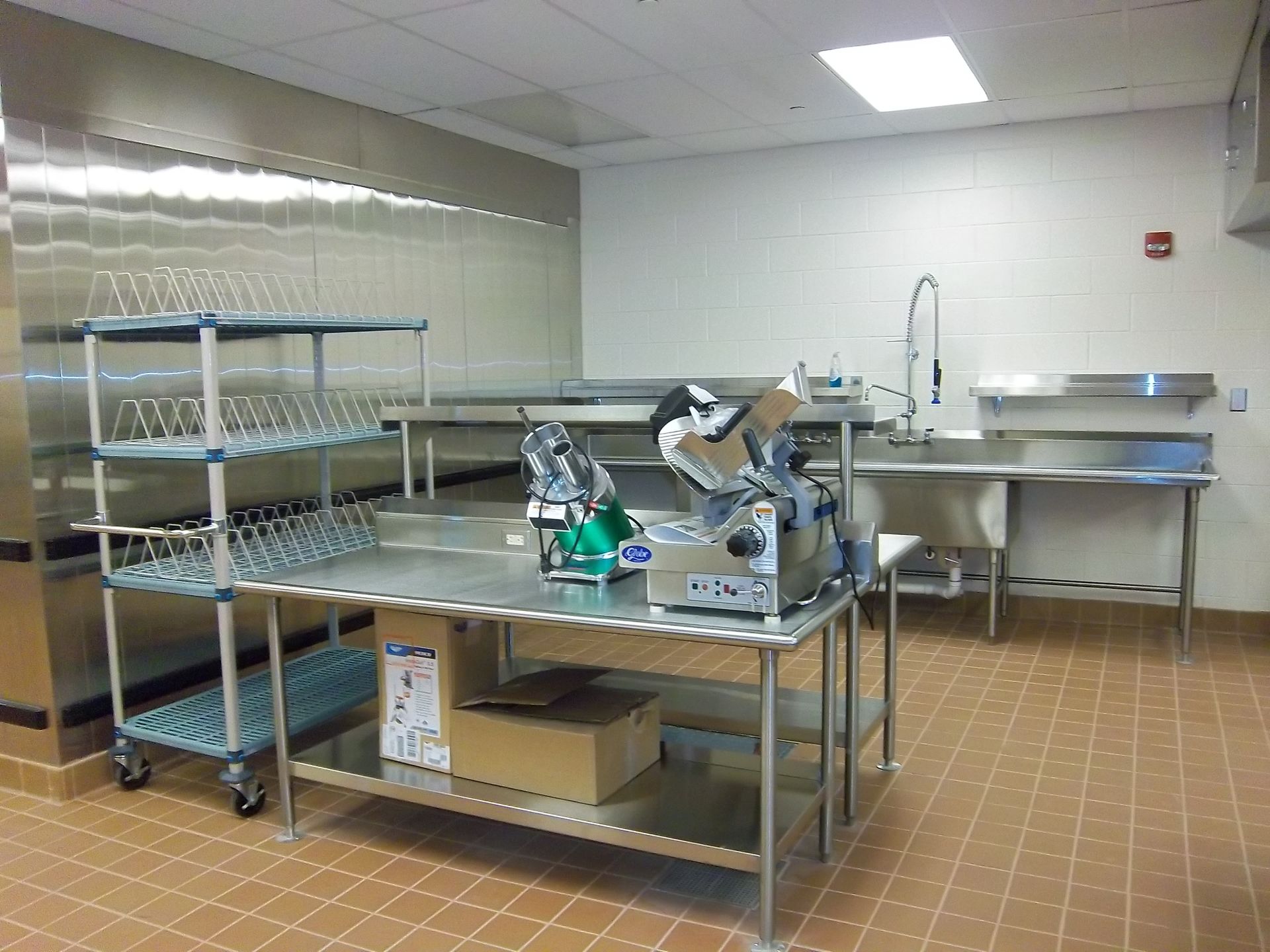 A commercial kitchen prep area with stainless steel tables, a rolling rack, and meat slicers on a tiled floor.