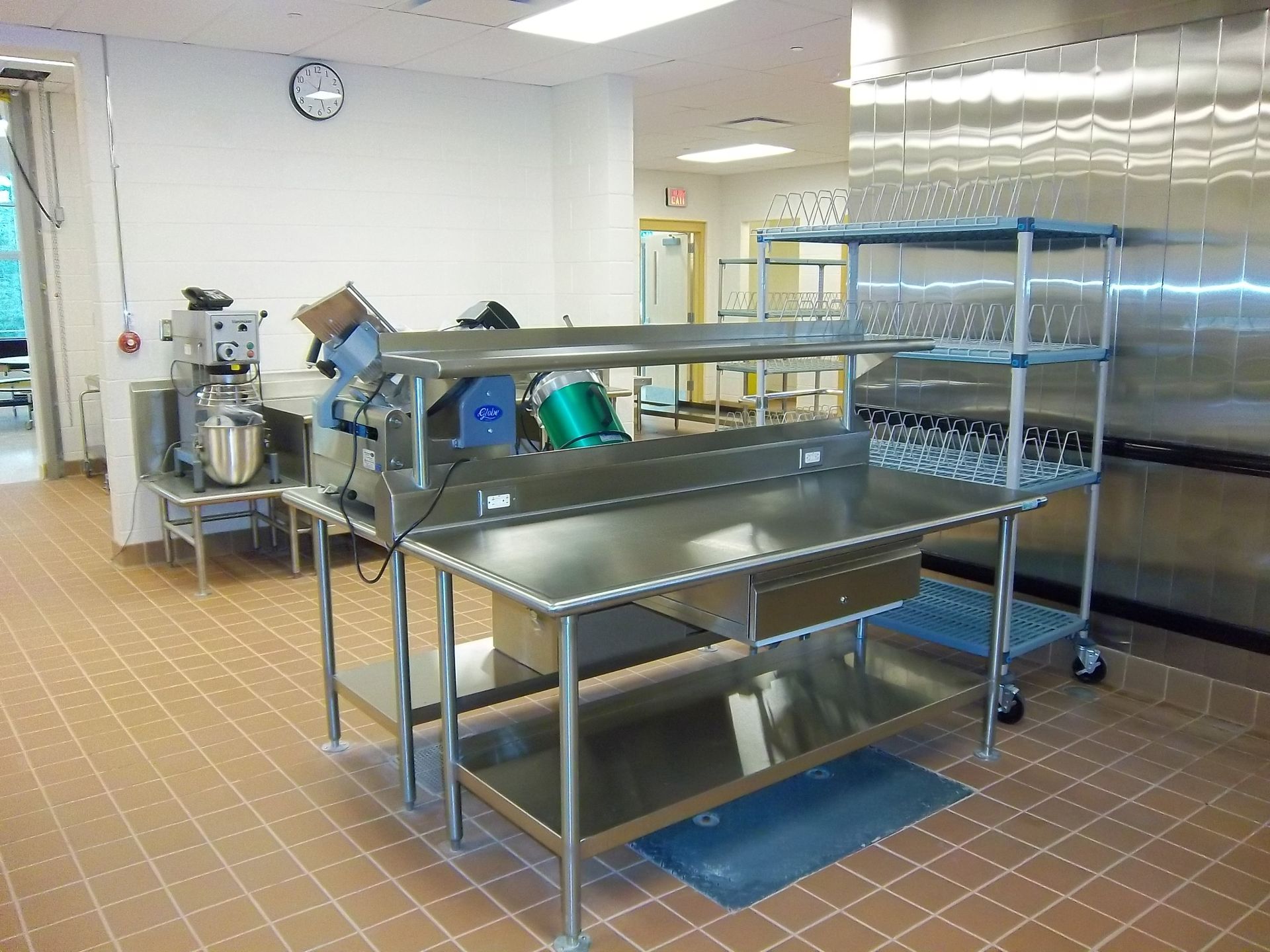 A commercial kitchen area featuring stainless steel prep tables, a food processor, and a metal shelving unit with glassware.