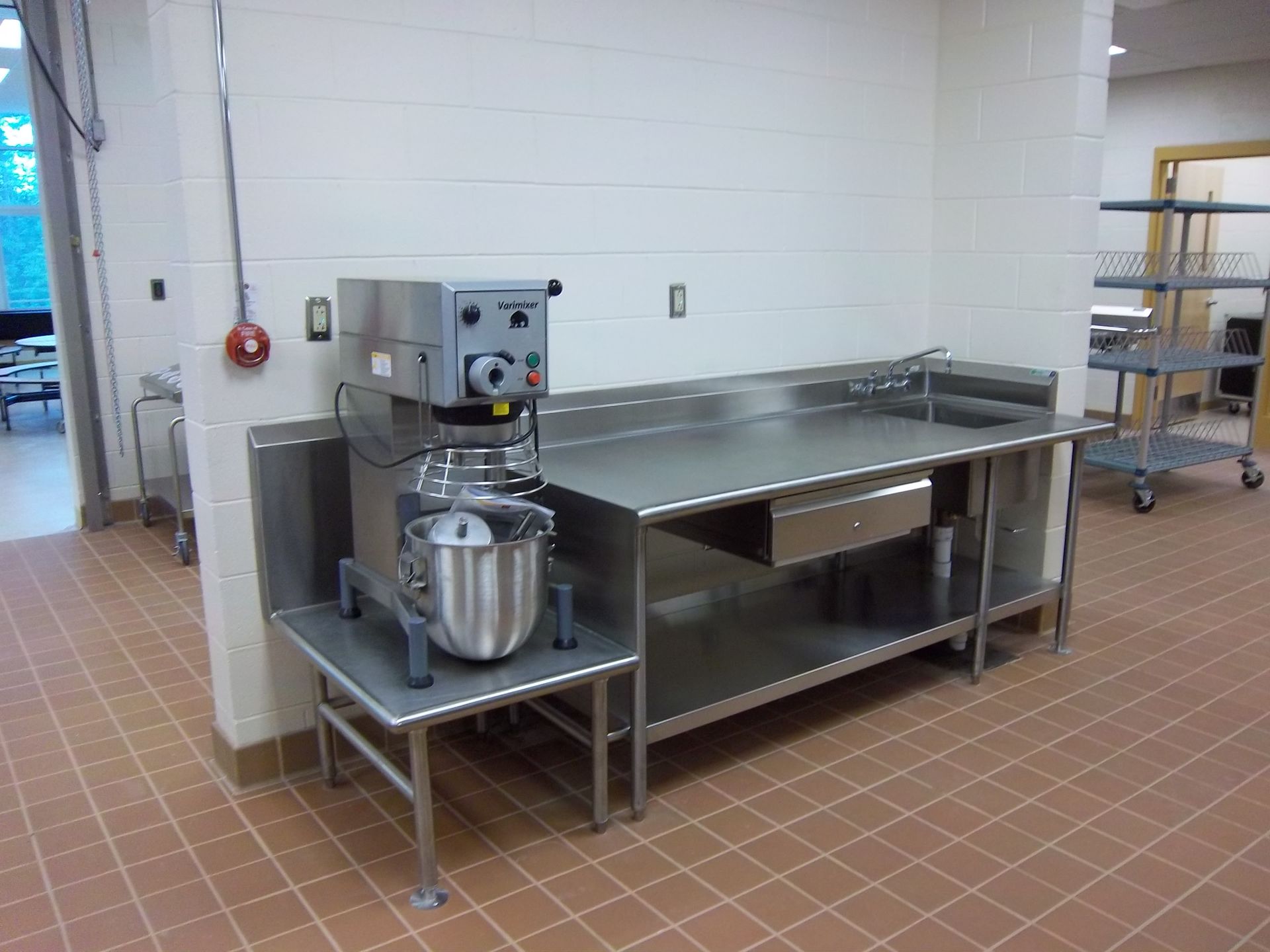 Industrial kitchen featuring a stand mixer on a small metal table next to a stainless steel prep station.