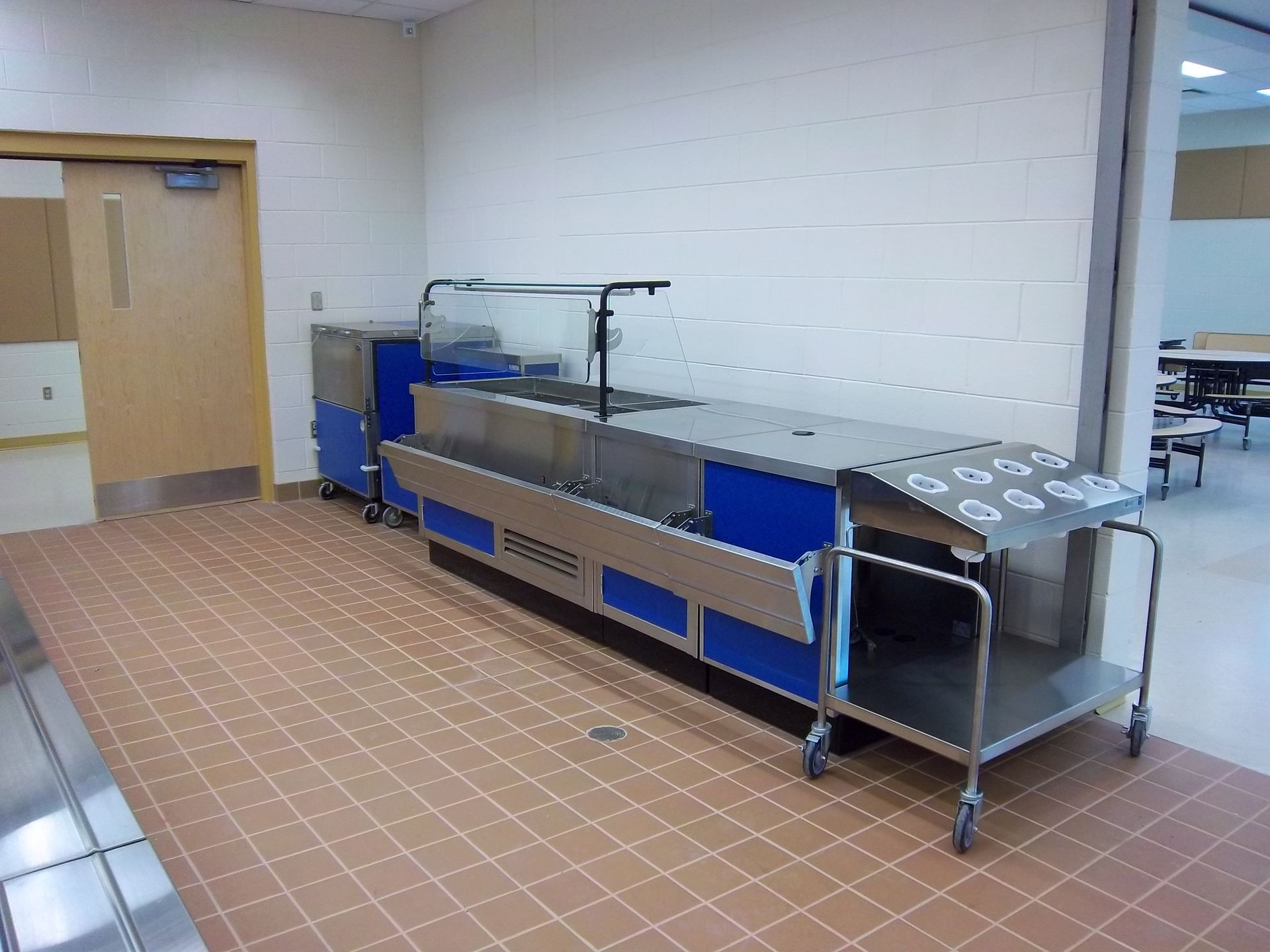 A long, stainless-steel food serving counter with blue panels in a room with tiled floors and a wooden door.