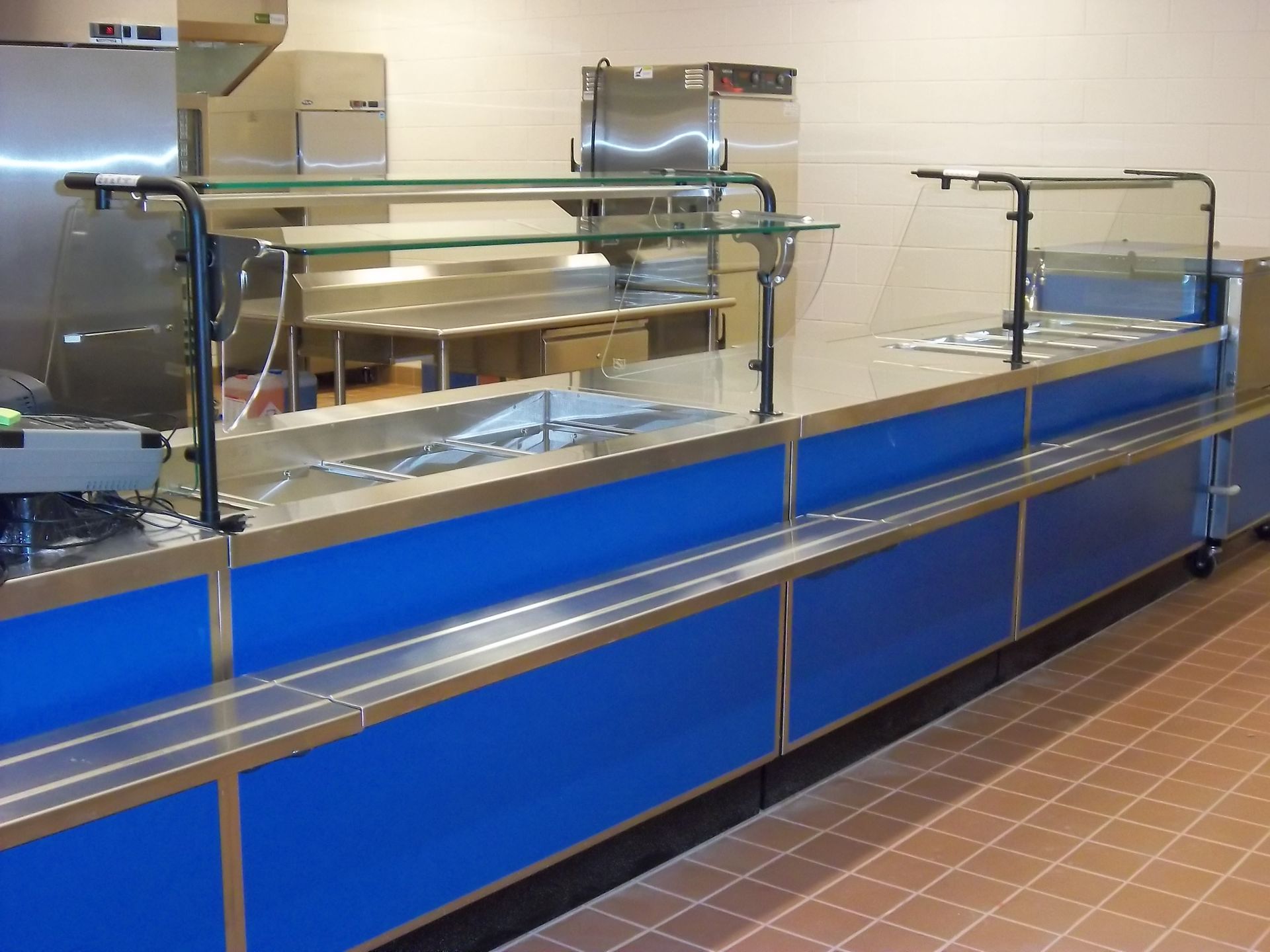 A blue stainless steel cafeteria serving line with a glass sneeze guard stands in a commercial kitchen with tiled floors.