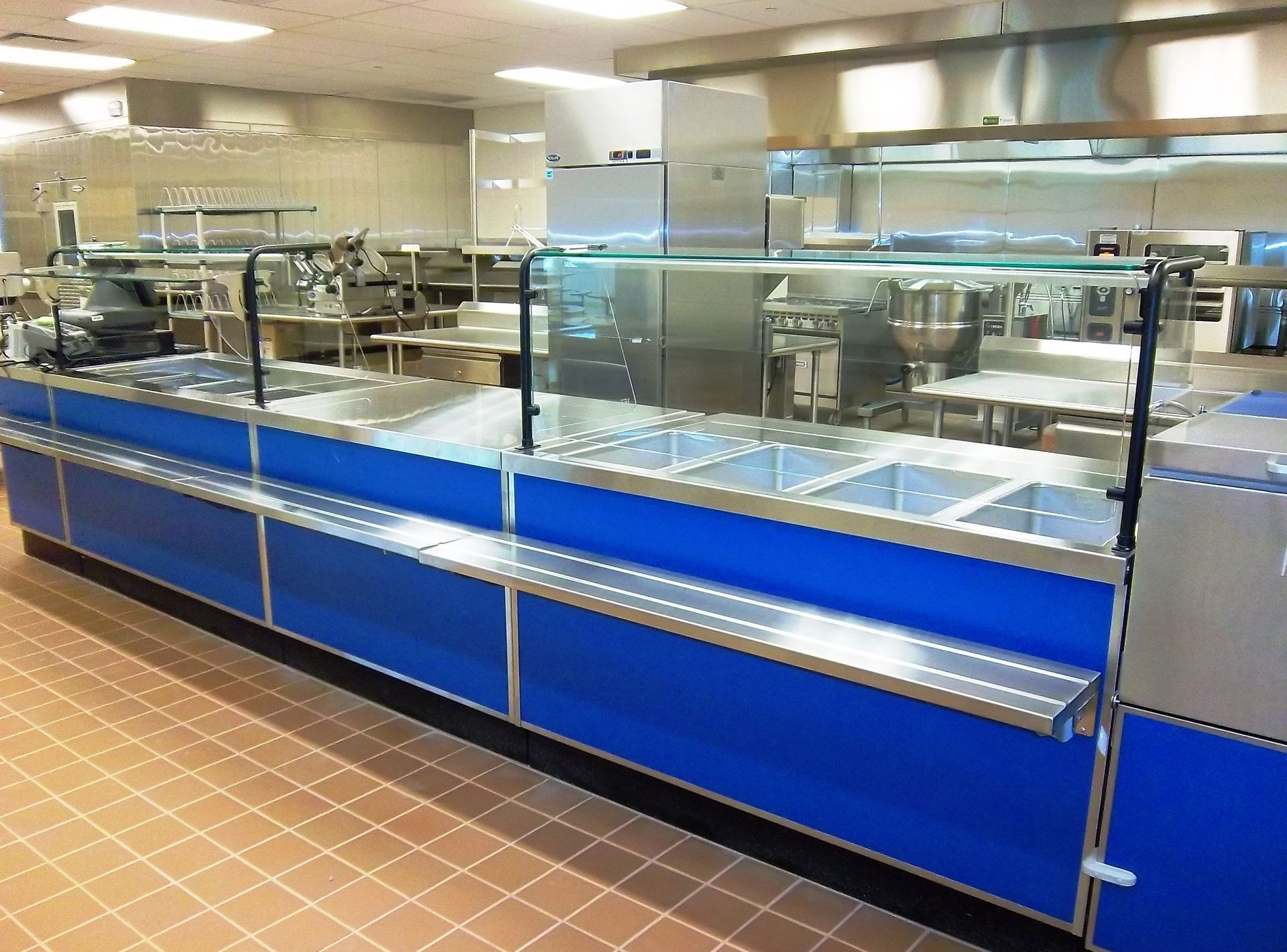 A commercial cafeteria serving line with a blue base, stainless steel top, and glass sneeze guards in a kitchen setting.