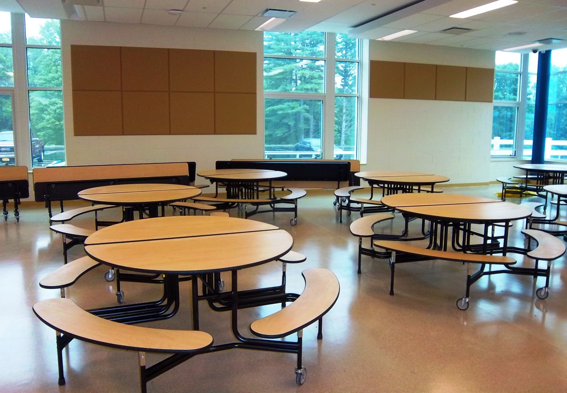 Empty school cafeteria with multiple round tables and attached curved benches under large windows.