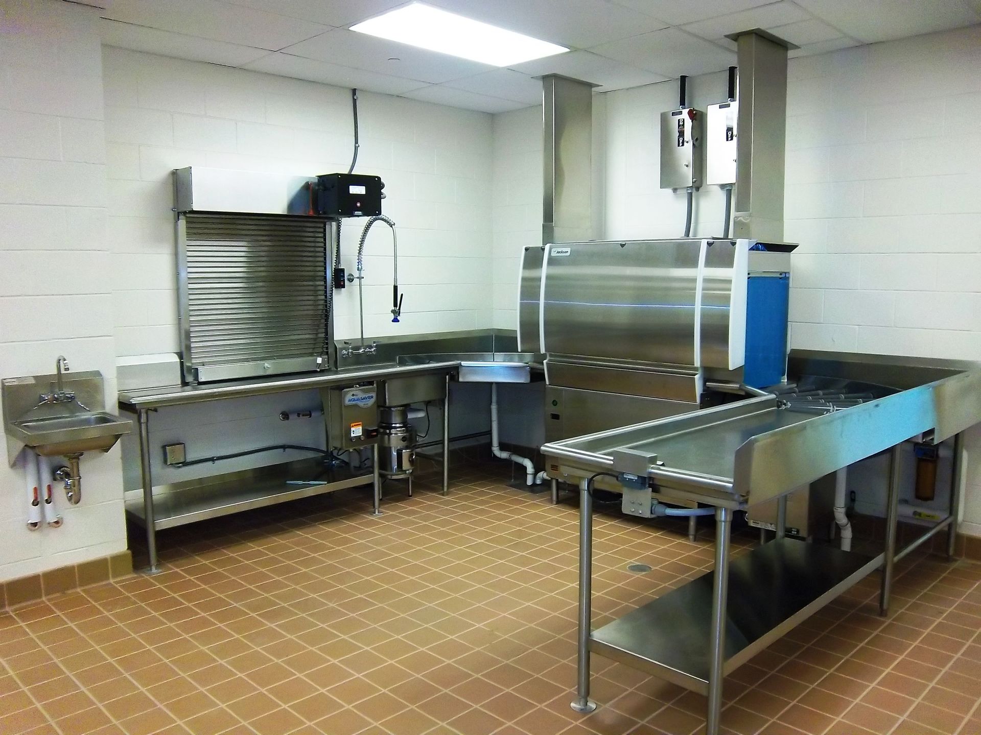 Industrial dishwashing station with a stainless steel sink, prep table, and large commercial dishwasher in a tiled room.