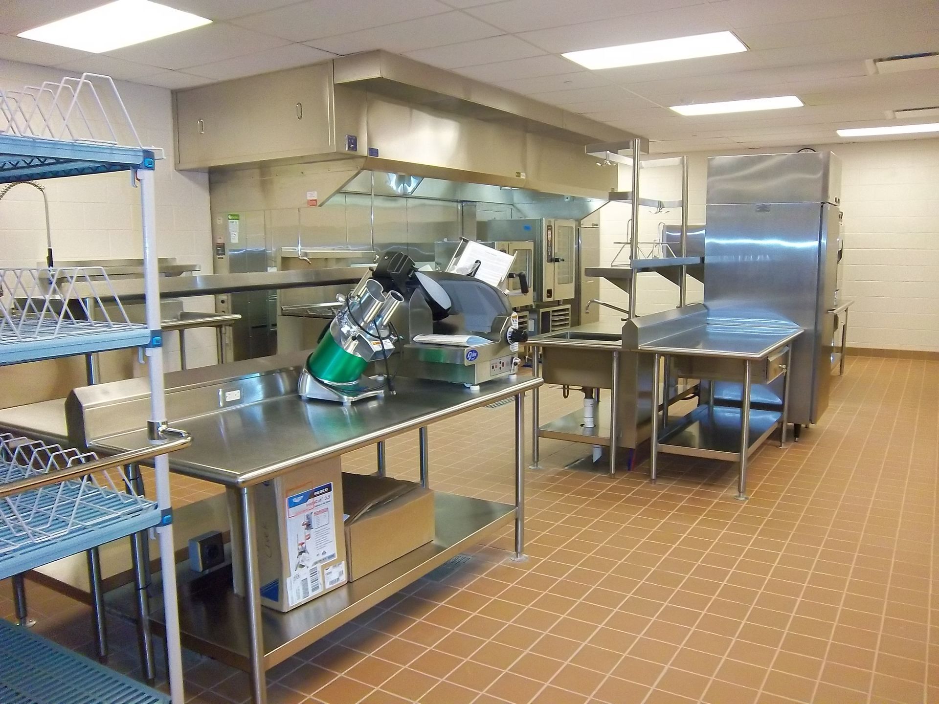 A commercial kitchen with stainless steel prep tables, a food slicer, and an industrial ventilation hood over the stove.