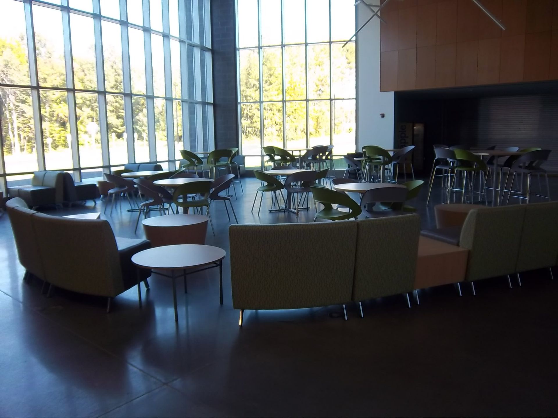 Modern lounge with floor-to-ceiling windows, green armchairs arranged in groups, and small tables on a dark floor.