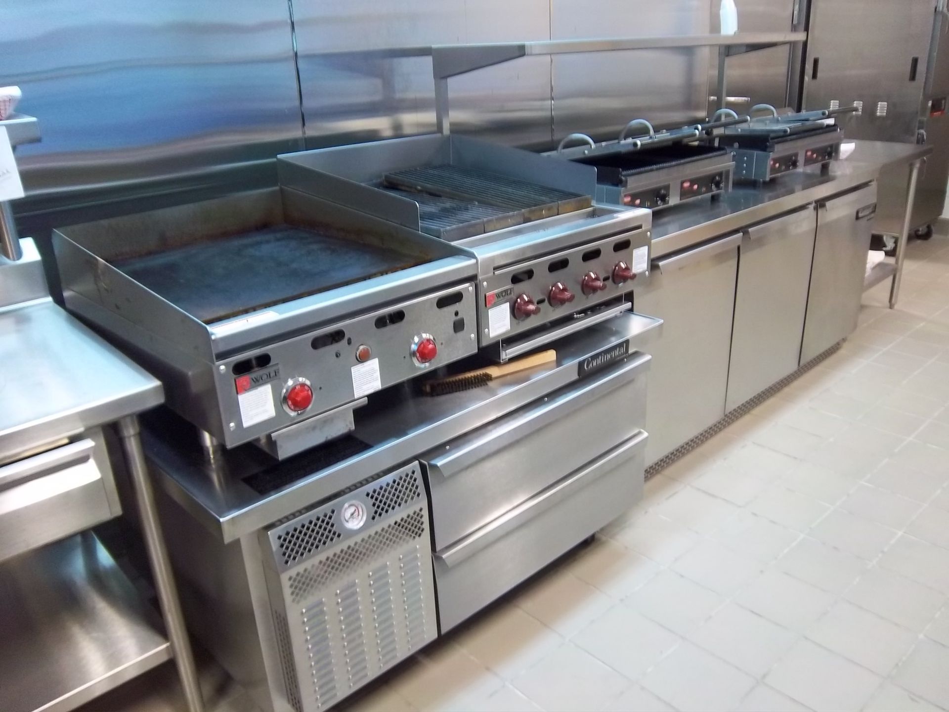Commercial kitchen stainless steel cooking line with a flat-top grill, charbroiler, and under-counter refrigeration units.