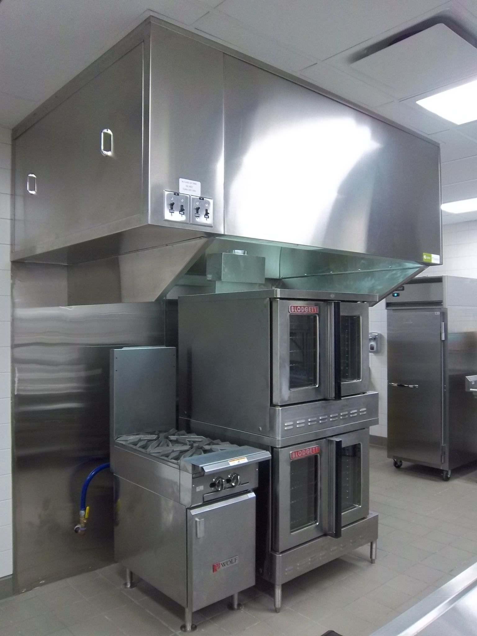 A commercial kitchen featuring stainless steel appliances, including a double-stack oven and a stove hood overhead.