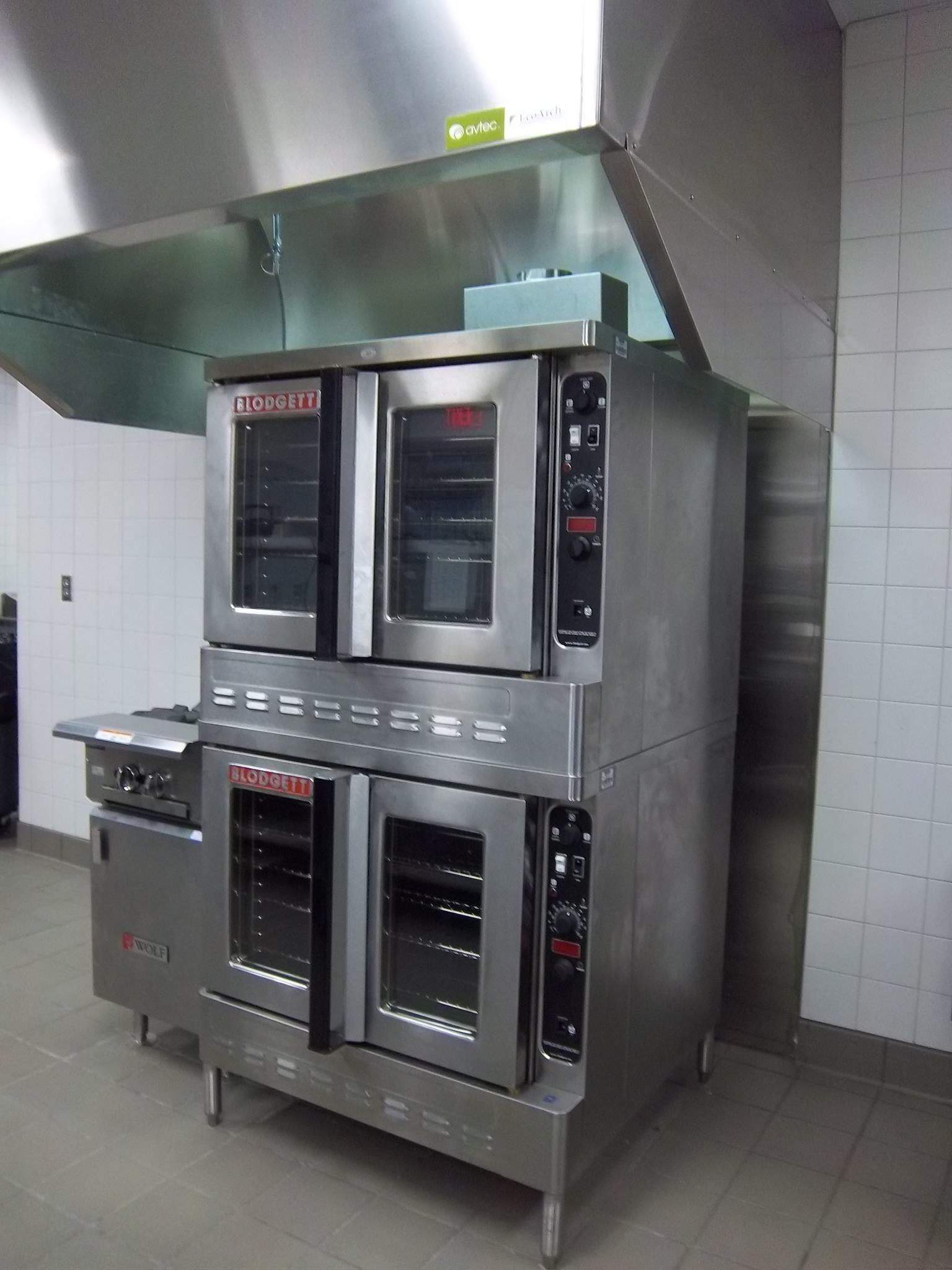 A commercial stainless steel double-stack convection oven unit situated under a metal ventilation hood in a kitchen.