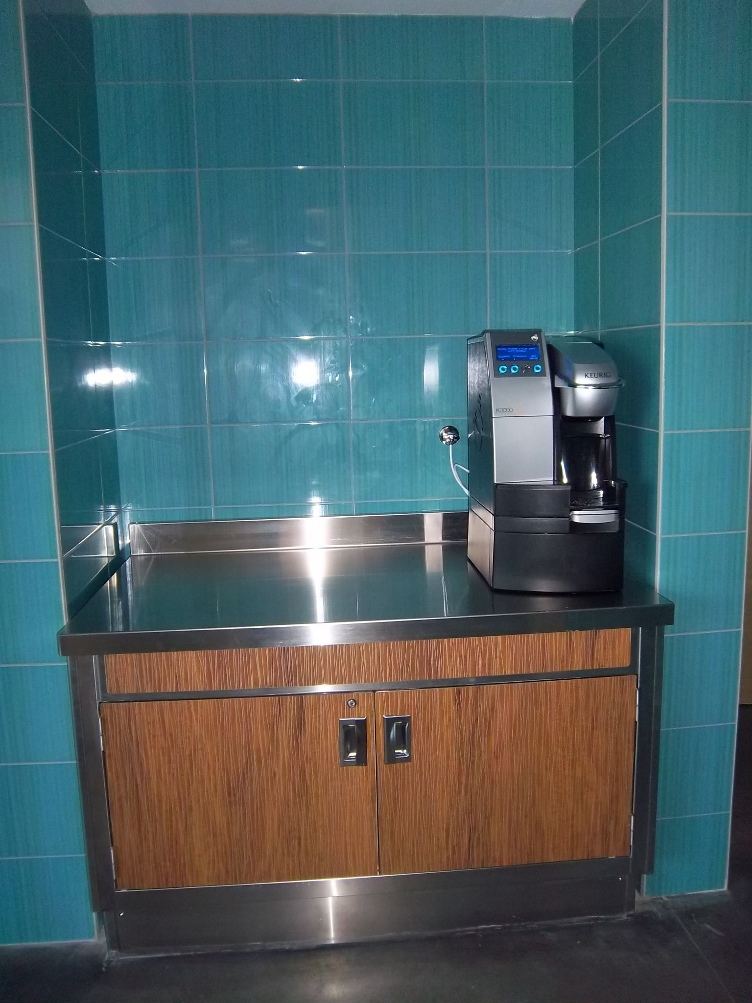 A black coffee machine sits on a stainless steel counter with wood-paneled cabinets against a teal tiled wall.