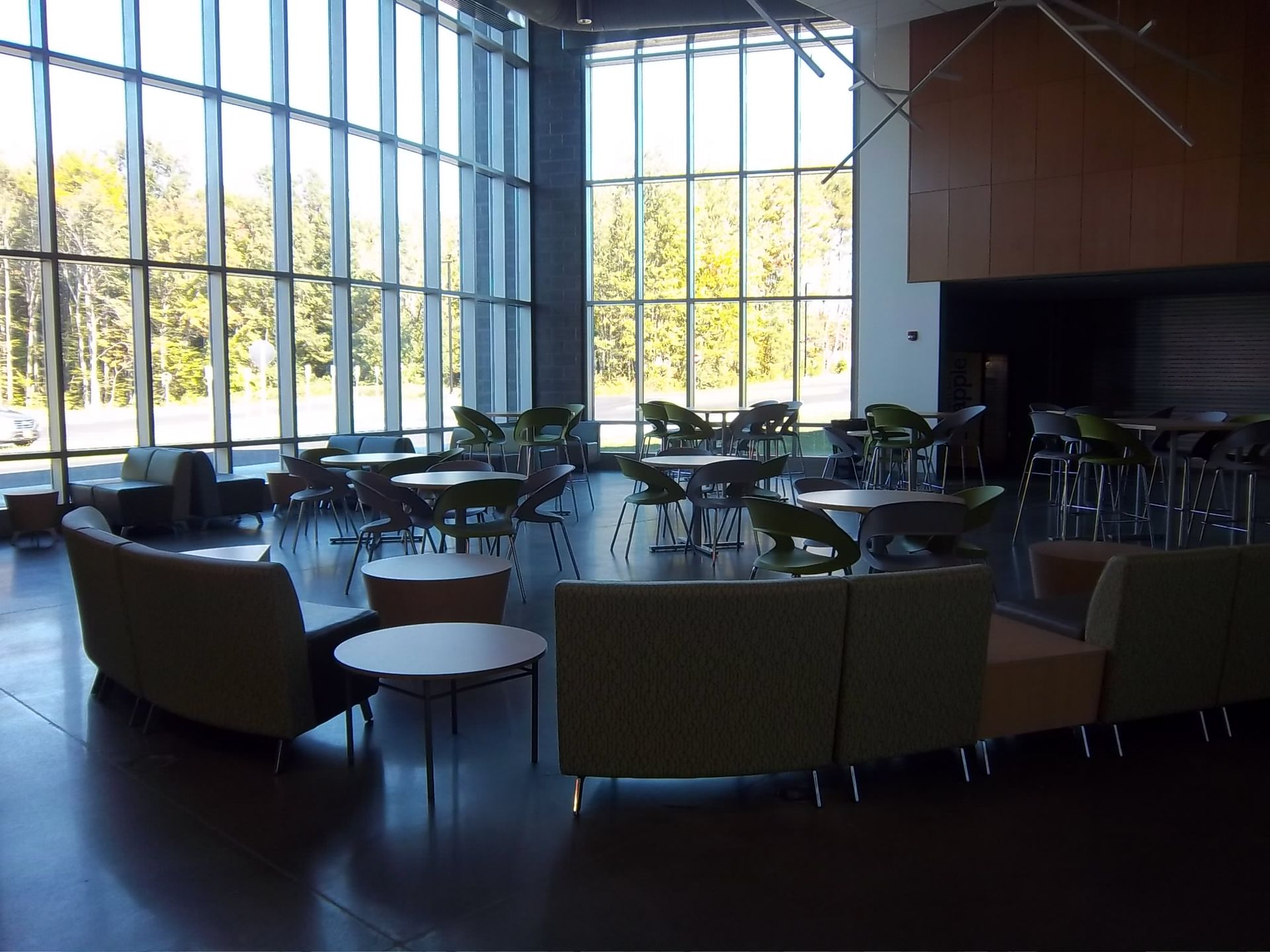 Modern lounge area with tall windows, arranged seating, round tables, and chairs on a polished floor in a bright atrium.
