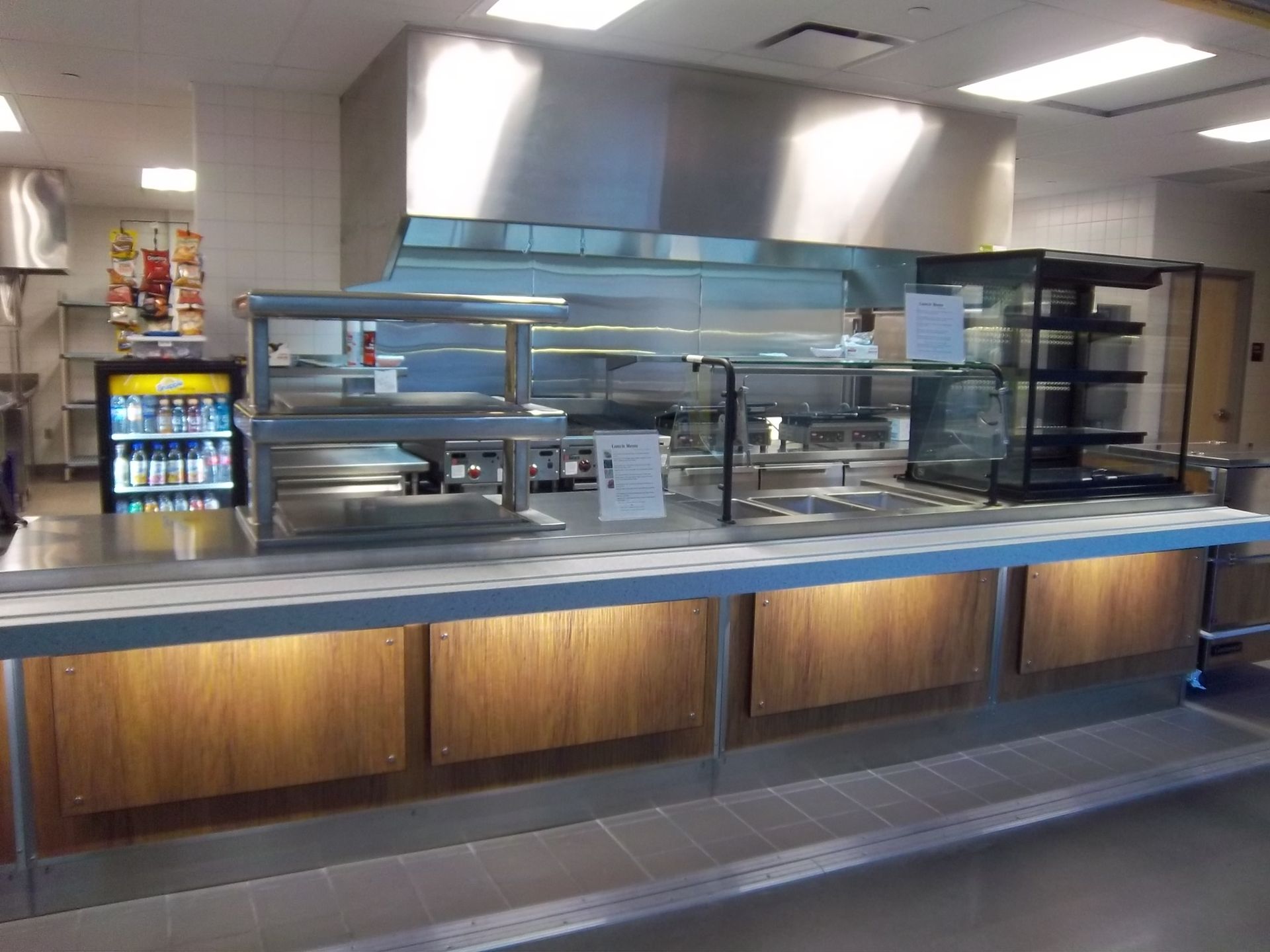 A stainless steel food service counter with wood panels and lit warming displays in a commercial kitchen setting.