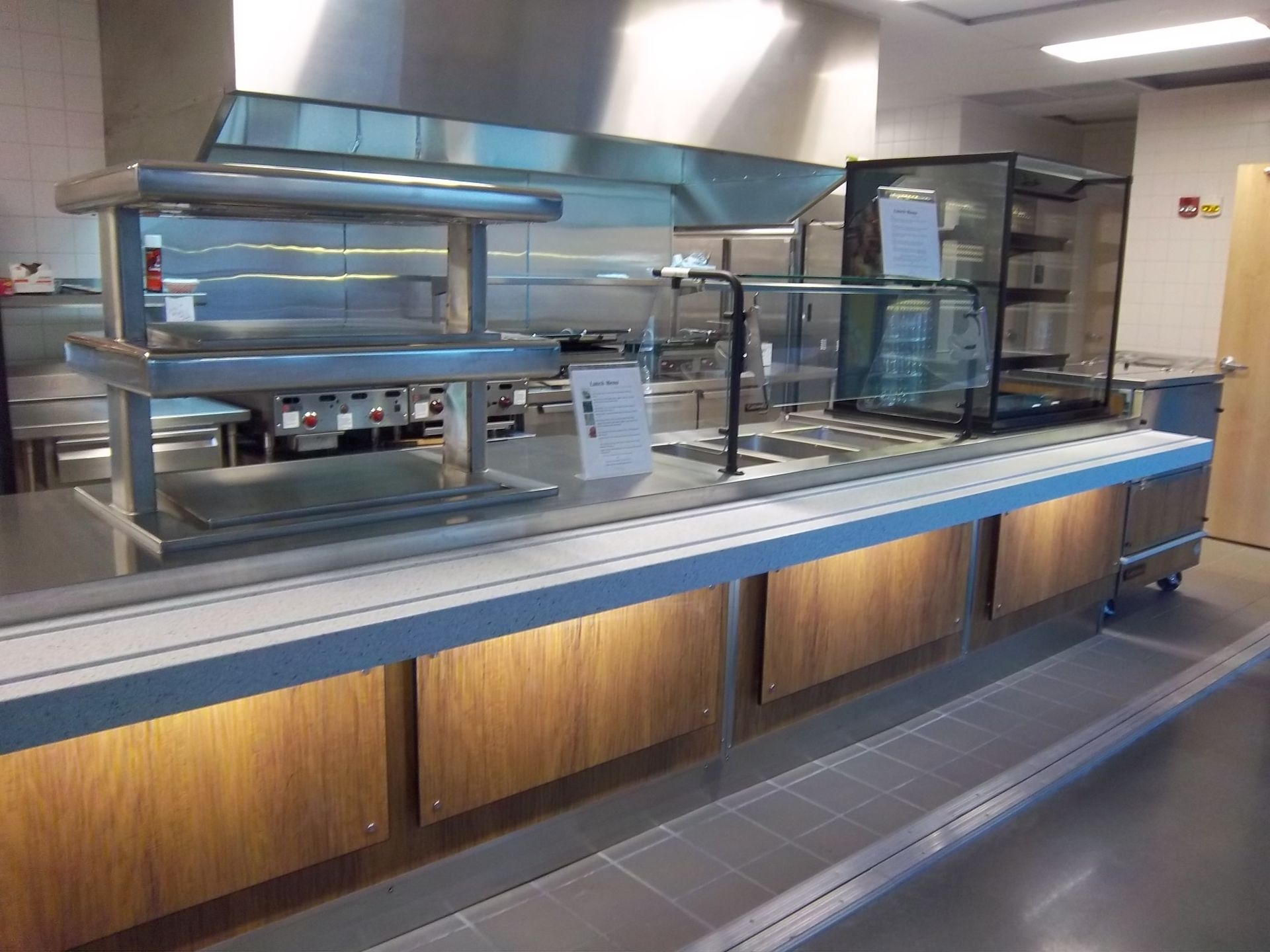A modern commercial cafeteria service counter with stainless steel equipment, glass sneeze guards, and wood paneling.