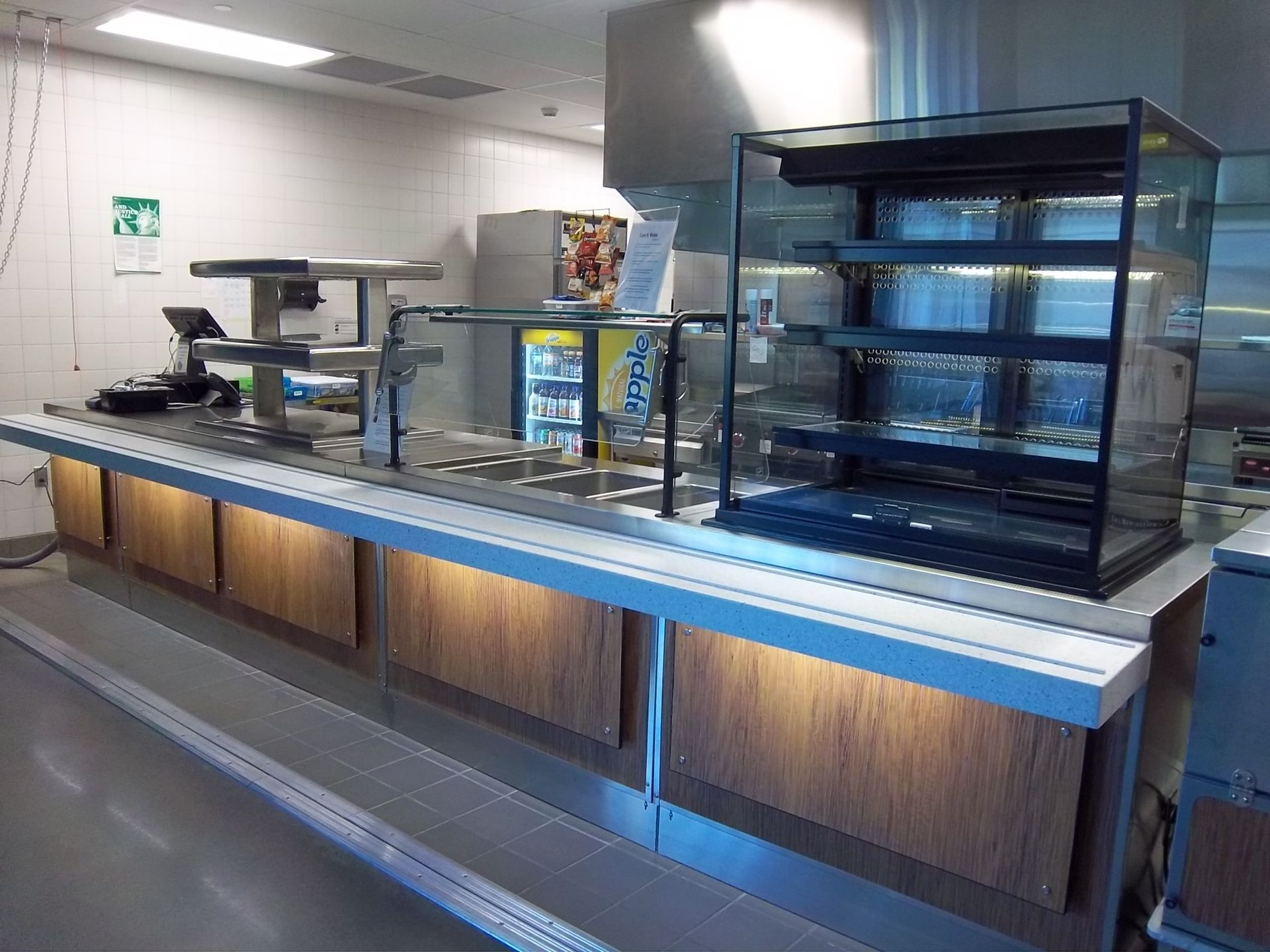 A cafeteria serving counter featuring a wood-paneled base with under-counter lighting, a glass display case, and equipment.