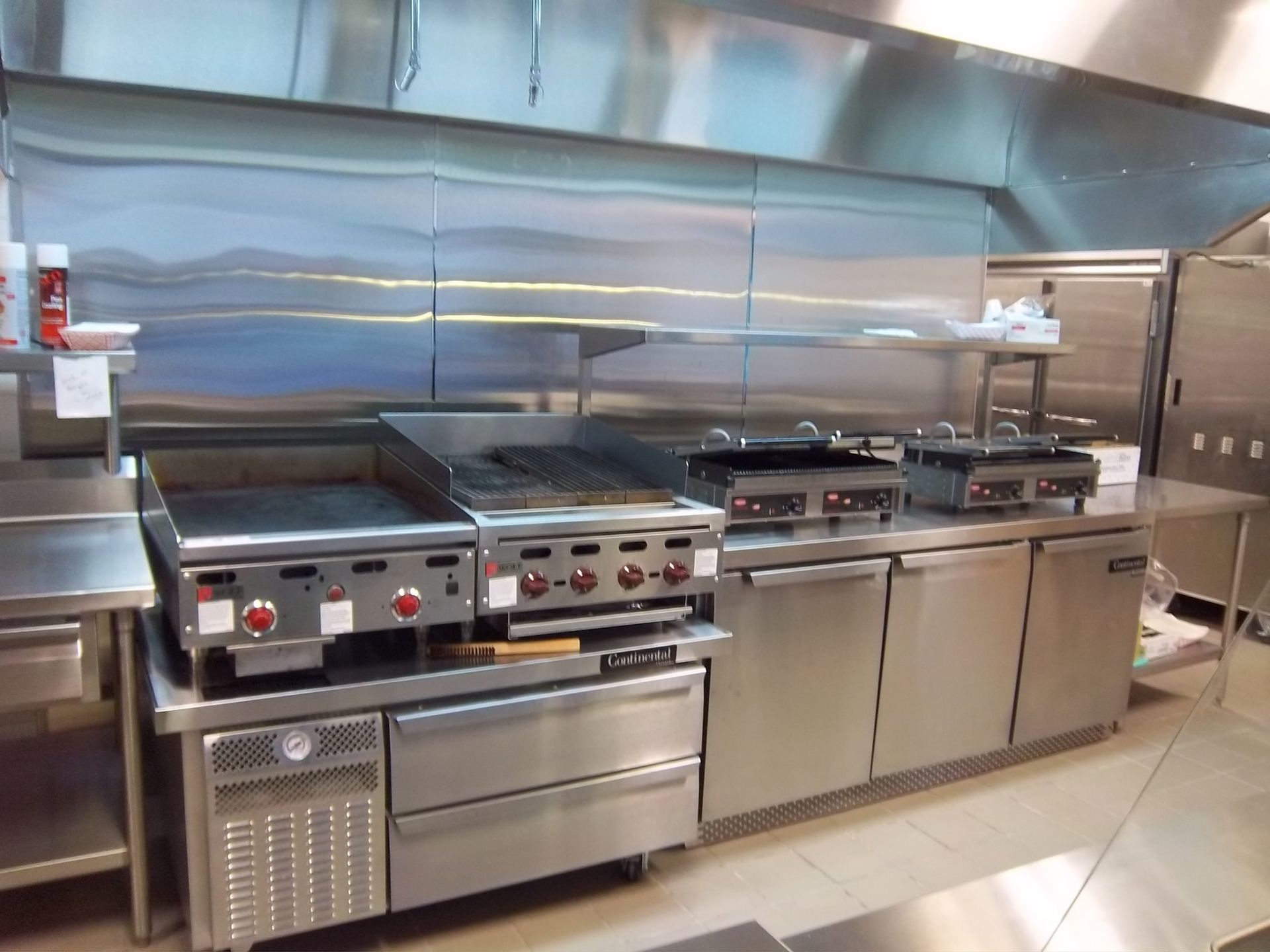 Stainless steel commercial kitchen with a flat-top grill, charbroiler, two panini presses, and under-counter refrigeration.