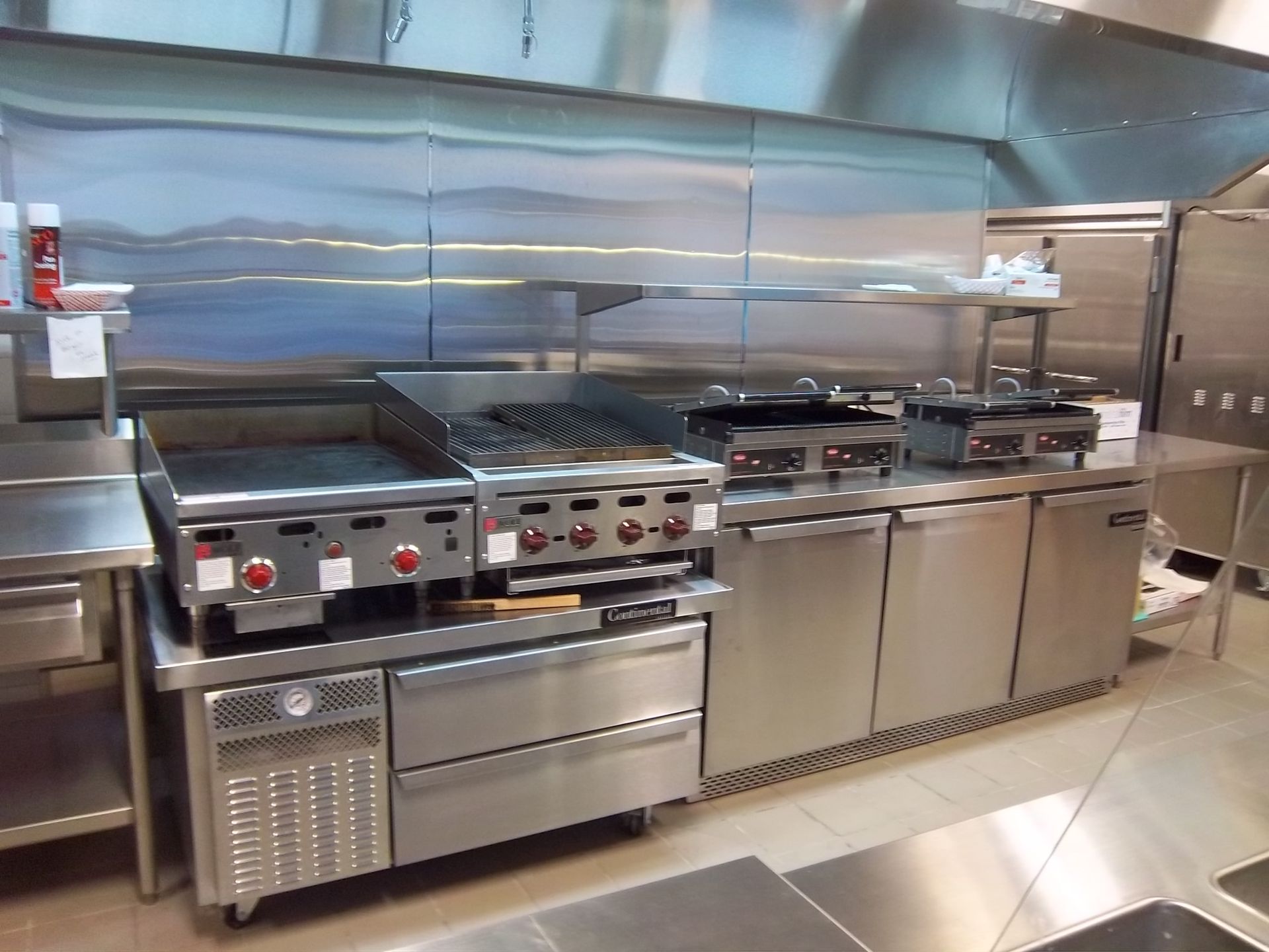 A stainless steel commercial kitchen line with a griddle, charbroiler, and under-counter refrigeration units.