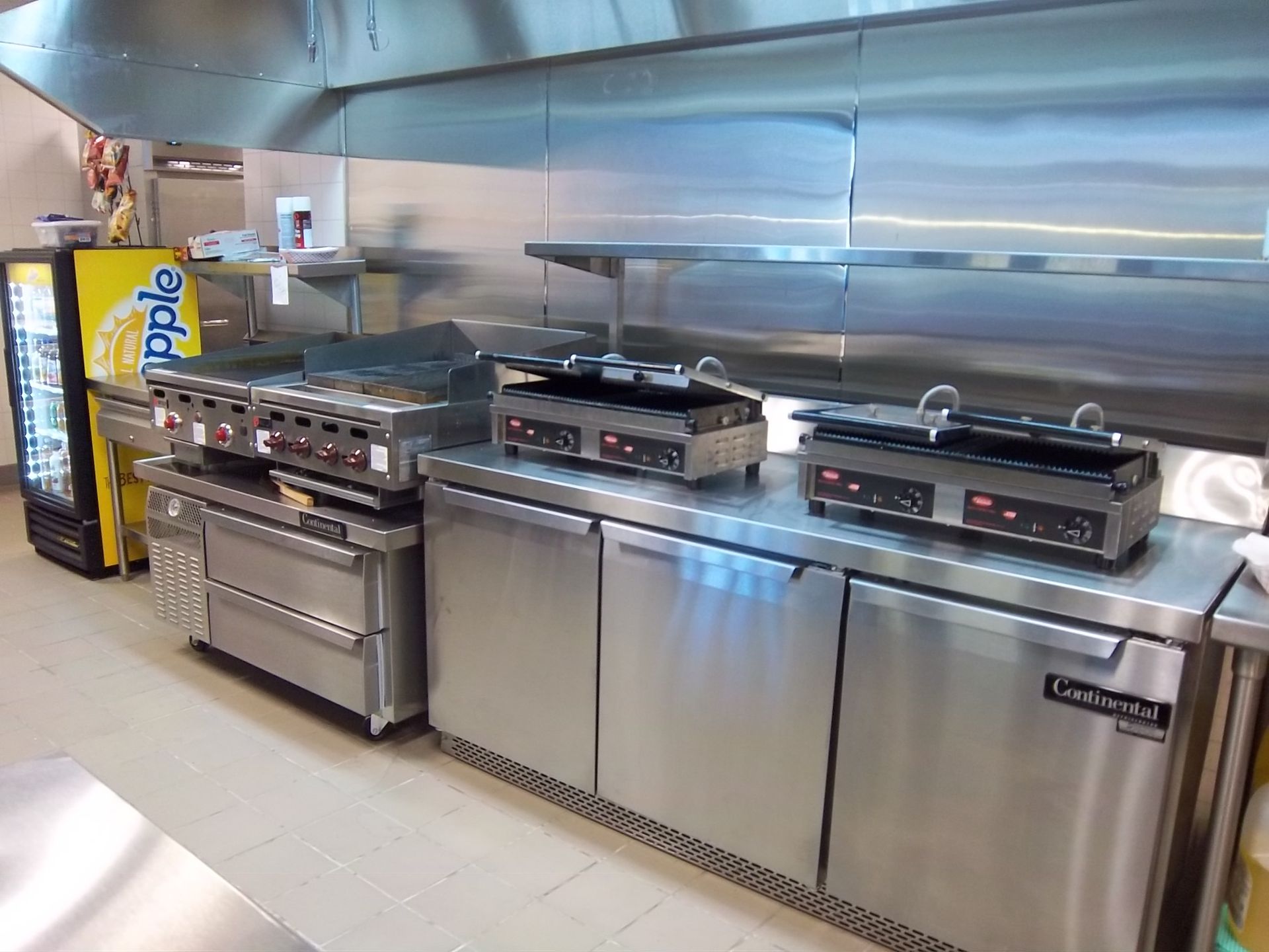 A commercial kitchen featuring a stainless steel prep station with two countertop grills beside a large gas griddle.