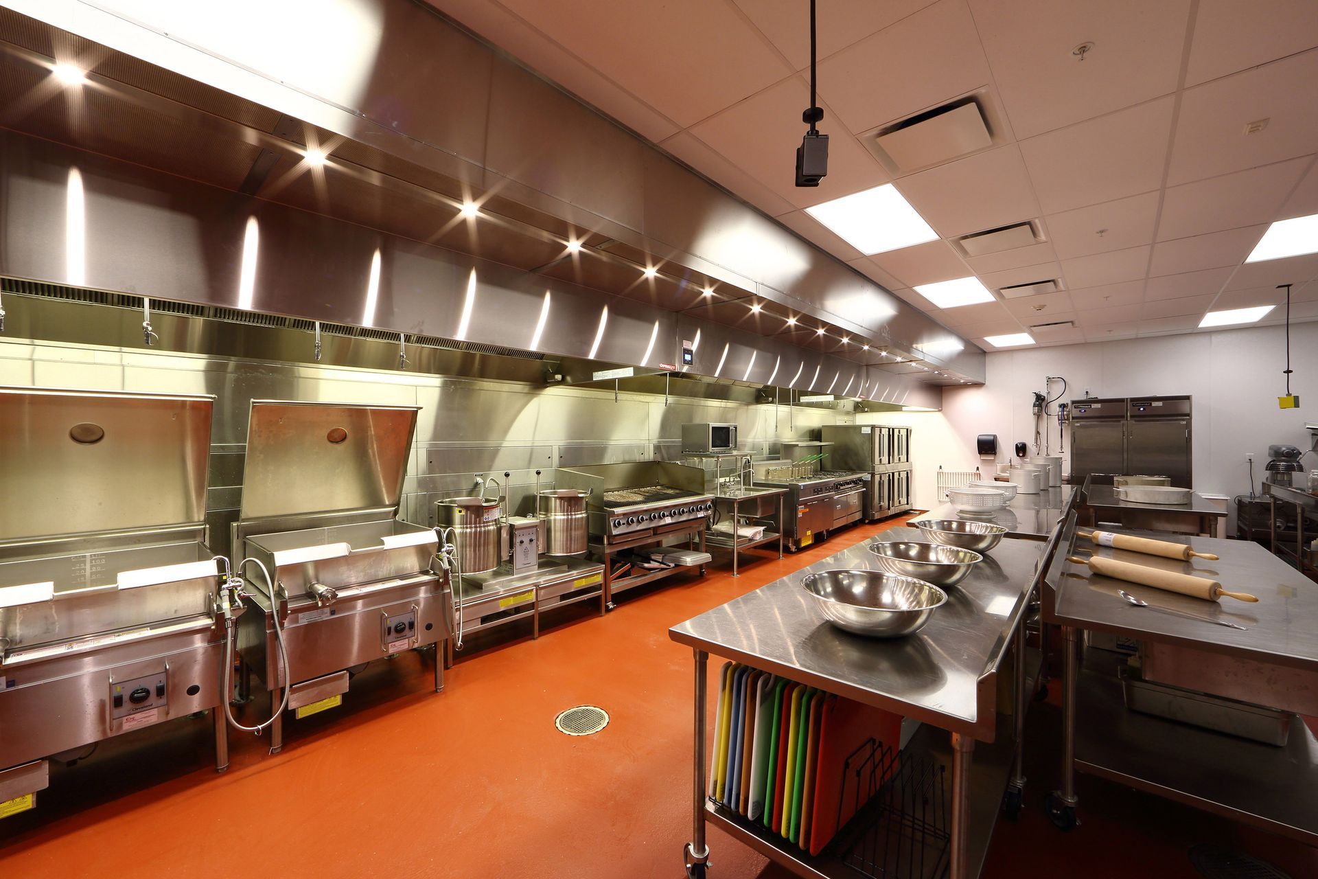 A clean, well-lit professional kitchen with stainless steel equipment, prep tables, and an orange floor.