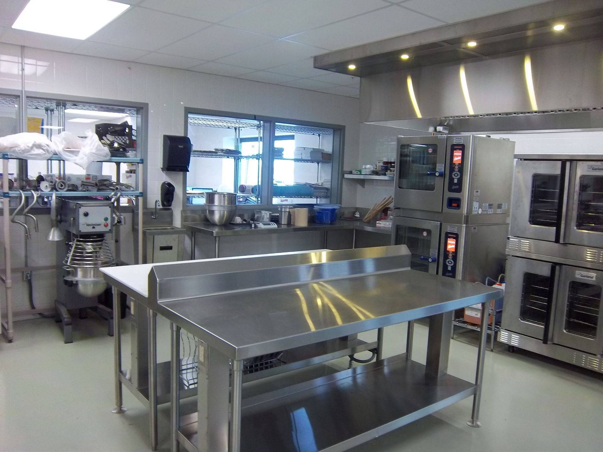 A bright, industrial kitchen with stainless steel prep tables, a large industrial mixer, and commercial ovens.