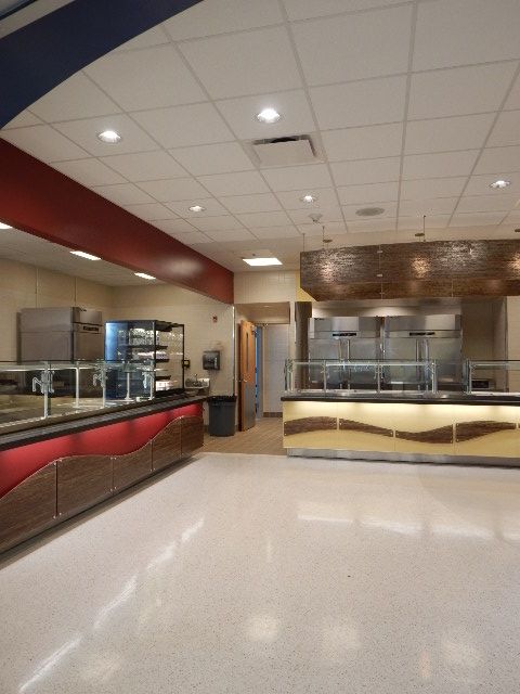 Modern cafeteria serving area with red and tan counters, food display cases, and bright overhead lighting.