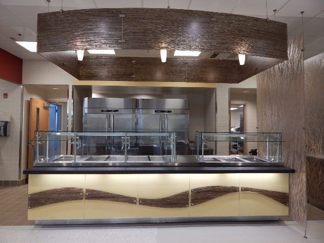 A commercial food serving station with glass sneeze guards, backlit counter panels, and an overhead wood-tone canopy.