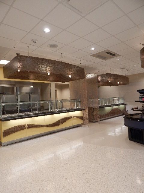 A cafeteria service area with two long serving counters, golden panels, and large textured light fixtures on a white floor.